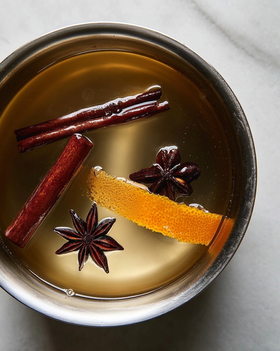 The image shows a close-up of a stainless steel pot with a clear liquid inside. Floating on the surface are two dark brown cinnamon sticks, two dark star anise pieces, and a thin piece of bright orange peel laying flat in the middle. The pot sits on a white marbled surface, and the spices and peel have a slightly shiny and wet texture from the liquid. The light reflects softly on the metal inside the pot, highlighting the rich colors and smooth texture of the ingredients photo taken with an iphone --ar 4:5 --v 7