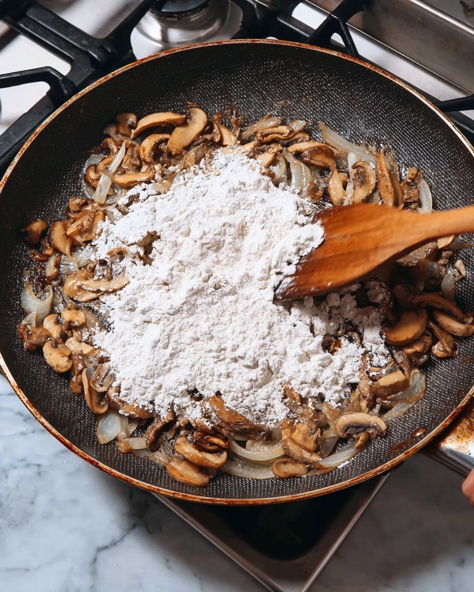 A close-up image of a frying pan on a stovetop, showing a layer of cooked, sliced mushrooms and onions that are light brown and soft. Over the mushrooms and onions, there is a thick layer of white flour sprinkled unevenly. A wooden spatula held by a woman's hand is seen on the right side, stirring the mixture. The frying pan has a dark textured surface with a metallic rim. The background includes parts of the stove with a white marbled surface beneath. Photo taken with an iphone --ar 4:5 --v 7