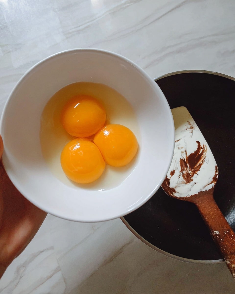 A close-up image shows a woman's hand holding a white bowl that contains three bright orange egg yolks. The bowl is tilted slightly over a dark pan, which has a white spatula with a brown handle resting inside it. The spatula shows some dark brown marks on its white part. The background surface has a white marbled texture. photo taken with an iphone --ar 4:5 --v 7