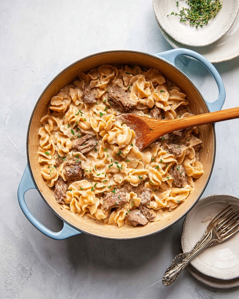 A light blue pot filled with creamy pasta and chunks of brown meat, mixed evenly in a light tan sauce with small green herb pieces on top, resting on a white marbled surface with a wooden spoon partially inside the pot, next to an empty white bowl and silver fork photo taken with an iphone --ar 4:5 --v 7