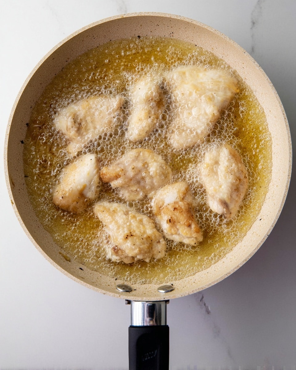 A light speckled beige pan filled with golden oil bubbles as six light cream-colored pieces of chicken are frying inside it. The chicken pieces show soft textures with some subtle brown spots beginning to appear. The pan handle is black with a silver connector, set against a white marbled surface. The image is taken from above, capturing the sizzling motion of the chicken cooking photo taken with an iphone --ar 4:5 --v 7