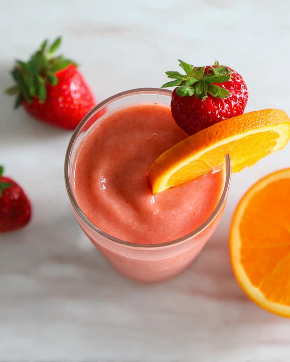 A clear glass filled with a smooth, thick pink smoothie is shown from above. The smoothie has a creamy texture and fills the glass almost to the top. On the rim, there is a bright red strawberry with green leaves and a fresh orange slice leaning against it. Nearby, on the white marbled surface, there is one whole strawberry with green leaves on the left side and a half orange on the right side. The colors are vibrant with the pink smoothie, red strawberry, and orange slice standing out against the light background photo taken with an iphone --ar 4:5 --v 7