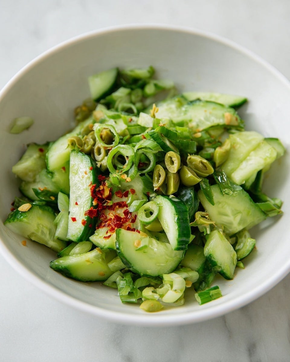 A white bowl sits on a white marbled surface filled with a fresh green salad made of sliced cucumbers, light green chopped olives, and thin round slices of green onions layered on top. The cucumbers are cut into uneven chunks and rings showing their bright green skin and pale watery inside. The olives are finely chopped and scattered mainly on one side. A small amount of reddish chili flakes peek through the cucumbers and green onions, adding a hint of color contrast. The overall texture is fresh and crunchy, with scattered pieces showing soft and slightly rough edges. The photo was taken with an iphone --ar 4:5 --v 7