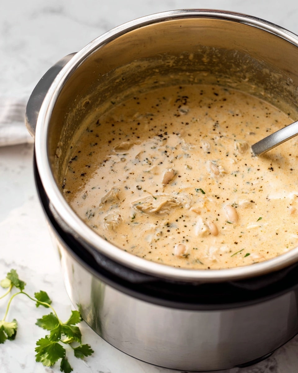 The image shows a creamy soup inside a stainless steel pot of an electric cooker, with a thick, light tan base speckled with small green herb pieces and black pepper. The soup has chunks of white meat and beans visible, all mixed in a smooth, rich texture filling most of the pot. The pot sits on a white marbled surface with a few green herb leaves scattered beside it. The lighting is soft and natural, highlighting the creamy texture and the shiny inside of the pot. Photo taken with an iphone --ar 4:5 --v 7