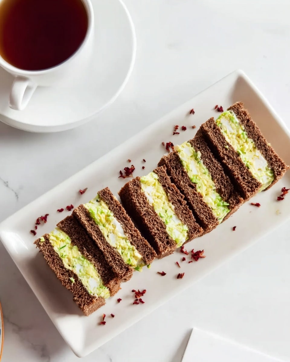 Five small rectangular sandwiches are neatly lined up on a long, white plate with a white marbled background. Each sandwich has three layers of dark brown bread with two layers of light green filling, slightly creamy with bits of white and green, resembling a vegetable or egg salad. Small red flakes are scattered around the plate for decoration. In the top left corner, a white cup holds a dark brown liquid, likely tea. The scene is bright and clean. photo taken with an iphone --ar 4:5 --v 7
