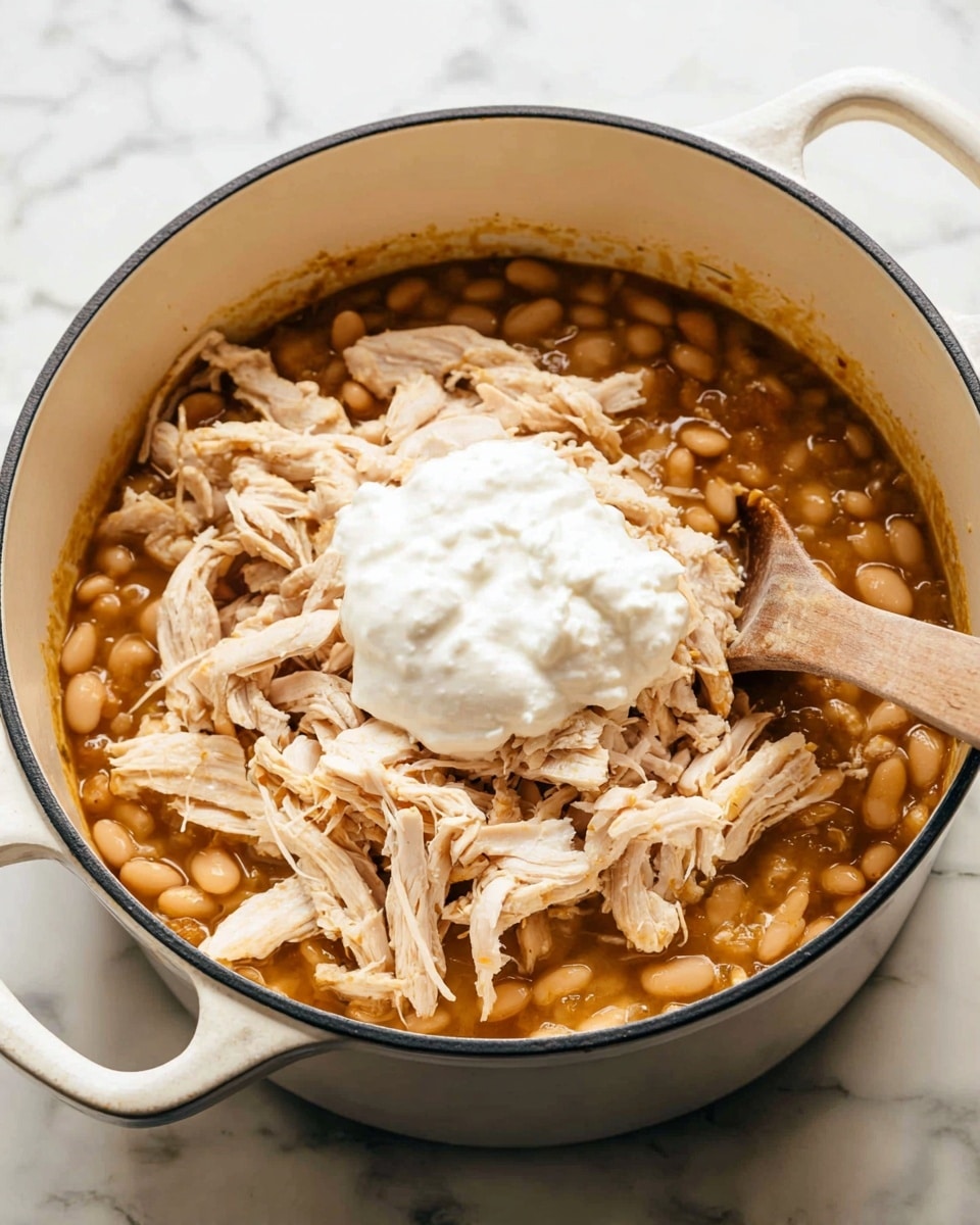 A large white pot contains a stew with three visible layers: the bottom layer shows a thick, brownish broth filled with soft white beans; on top of the beans, there are shredded pieces of light beige cooked chicken spread evenly; the top layer is a dollop of thick white cream sitting in the center of the chicken. A wooden spoon is placed inside the pot, resting on the stew. The pot is on a white marbled surface. Photo taken with an iphone --ar 4:5 --v 7