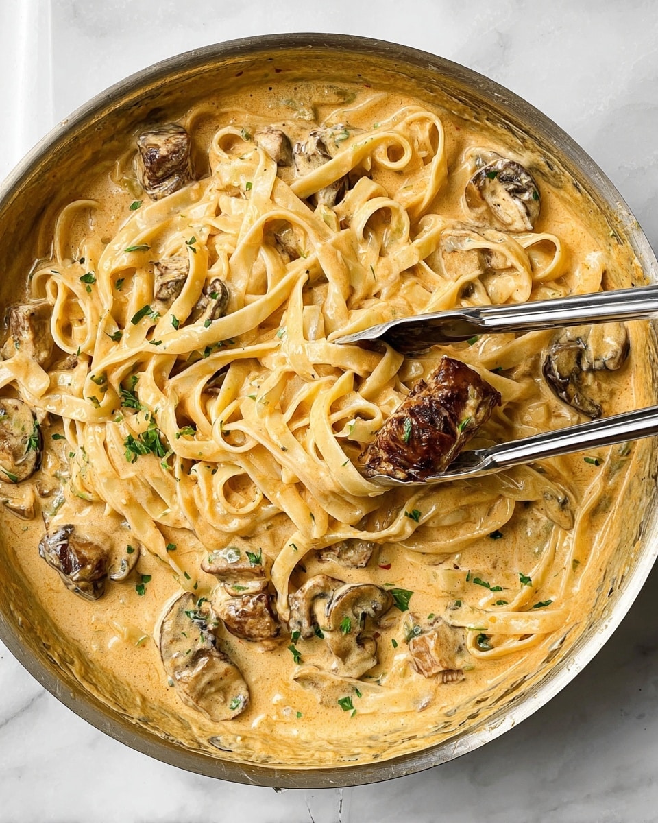 This image shows a close-up of a creamy pasta dish in a pan. There are thick flat noodles, light beige in color, fully coated in a smooth, creamy sauce that is a warm light orange shade with specks of herbs. Mixed in are several large chunks of browned mushrooms that add texture and dark brown spots. Small green herb pieces are scattered on top of the sauce. Two silver tongs hold some noodles and a mushroom piece in the center, lifting them slightly. The pan sits on a white marbled surface. photo taken with an iphone --ar 4:5 --v 7