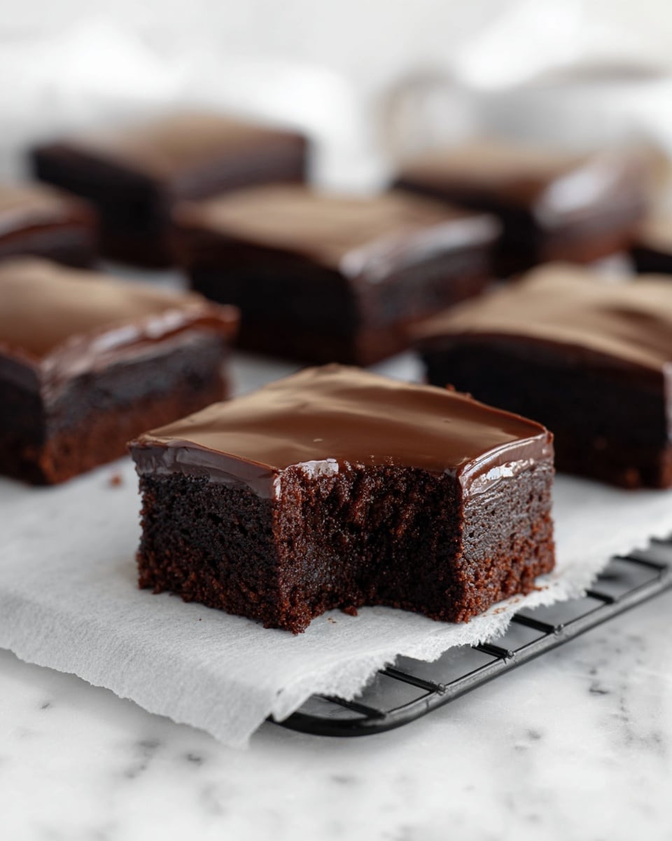 The image shows a close-up of a square chocolate brownie with a bite taken from one corner, revealing its moist and dense dark brown interior. The brownie has two layers: a thick, rich dark brown base with a rough texture, and a smooth, shiny chocolate glaze on top that covers the entire surface evenly. In the background, more brownies of the same kind are arranged on a sheet of white parchment paper, all placed on a black rack. The scene is set against a white marbled surface, with soft lighting that highlights the rich colors and texture of the brownies. Photo taken with an iphone --ar 4:5 --v 7