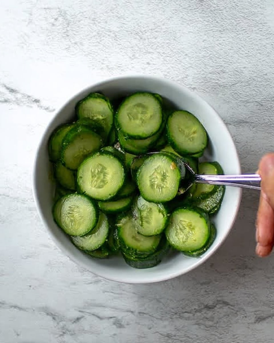 A white bowl filled with many slices of dark green cucumber, each slice showing the lighter green inside with seeds, placed on a white marbled surface. A woman's hand holds a silver spoon inside the bowl, lightly touching some cucumber slices. Photo taken with an iphone --ar 4:5 --v 7