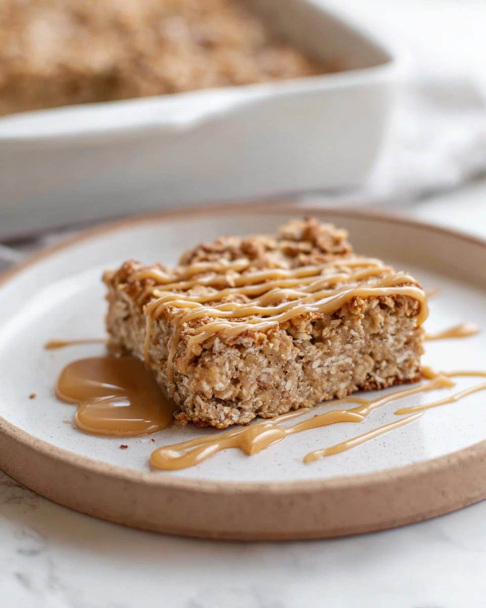 A square-shaped baked oatmeal bar sits on a round white plate with a white marbled surface underneath. The bar has a thick bottom layer with a rough, crumbly texture in light beige and brown tones. On top, there is a drizzle of creamy caramel-colored sauce that adds a smooth contrast to the coarse oatmeal. The plate has a slight shine, and some sauce drips loosely around the bar's edges. In the background, a blurred white baking dish with more oatmeal bars is visible. Photo taken with an iphone --ar 4:5 --v 7