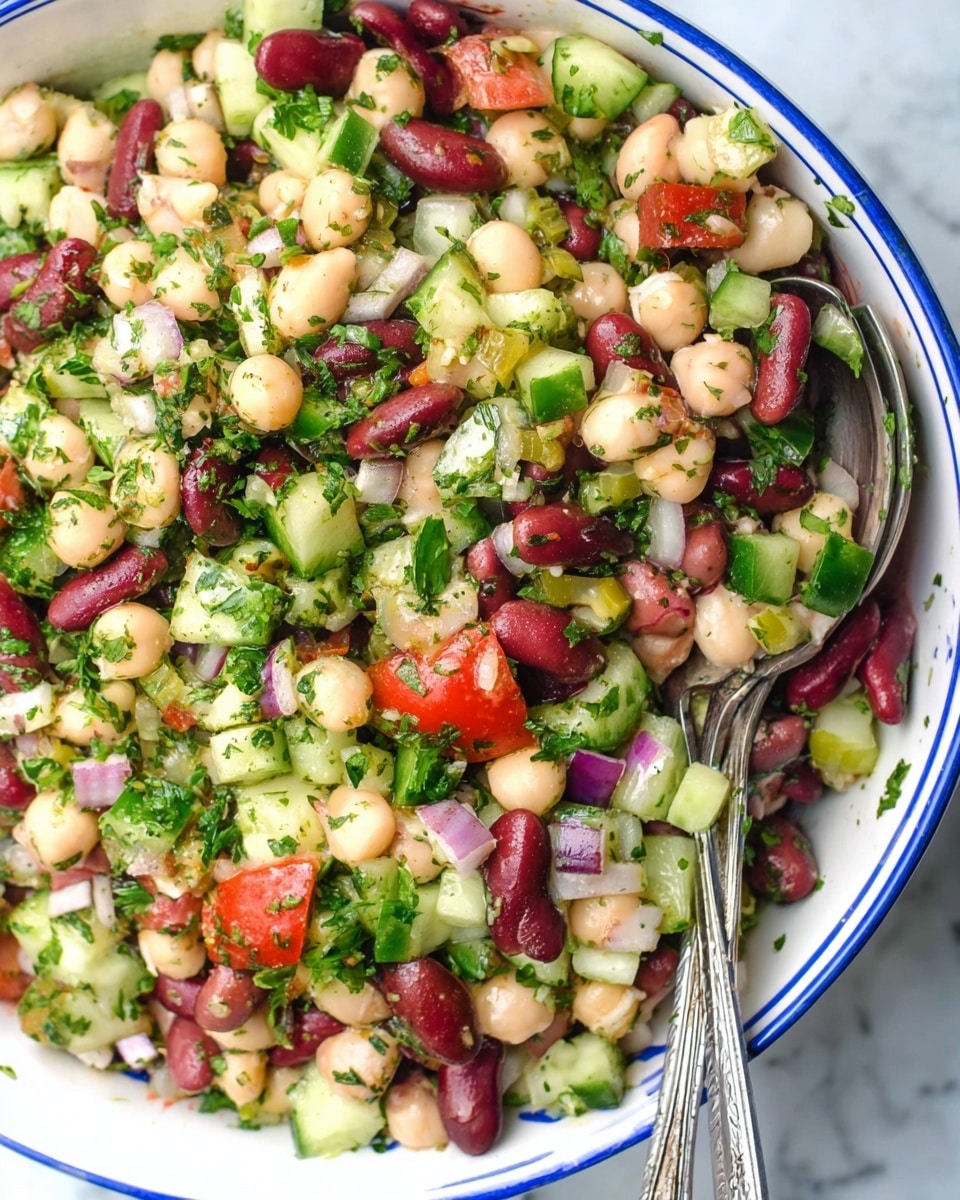 A close-up view of a colorful bean salad in a white bowl with a blue rim, filled with three layers of mixed ingredients: large red kidney beans, creamy chickpeas, and pale white beans forming the base layer, diced green cucumbers and chopped fresh green herbs scattered on top, with finely chopped red onion and bright red tomato pieces adding bursts of color, all mixed together and sprinkled with dried green herbs, two silver serving spoons resting inside on a white marbled surface photo taken with an iphone --ar 4:5 --v 7