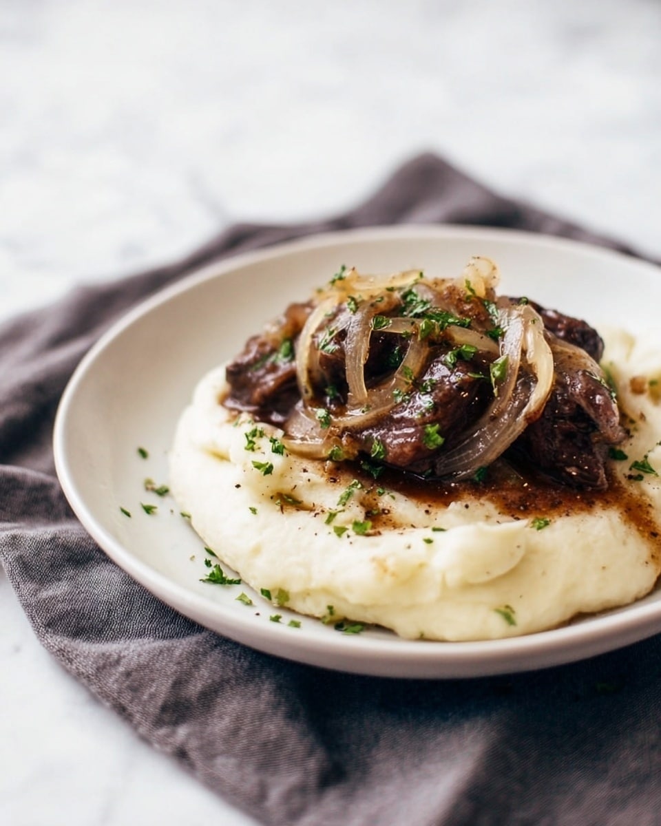 The dish shows a white plate with two main layers: the bottom layer is smooth, creamy mashed potatoes in a circular shape, light off-white in color with a soft texture. On top there are pieces of dark brown meat covered with a glossy brown sauce, mixed with thin slices of cooked onions that are light golden and translucent. The meat and onions have small bright green herb pieces sprinkled over them adding color. The plate is placed on a soft dark gray cloth, while the background is a white marbled texture. Photo taken with an iphone --ar 4:5 --v 7