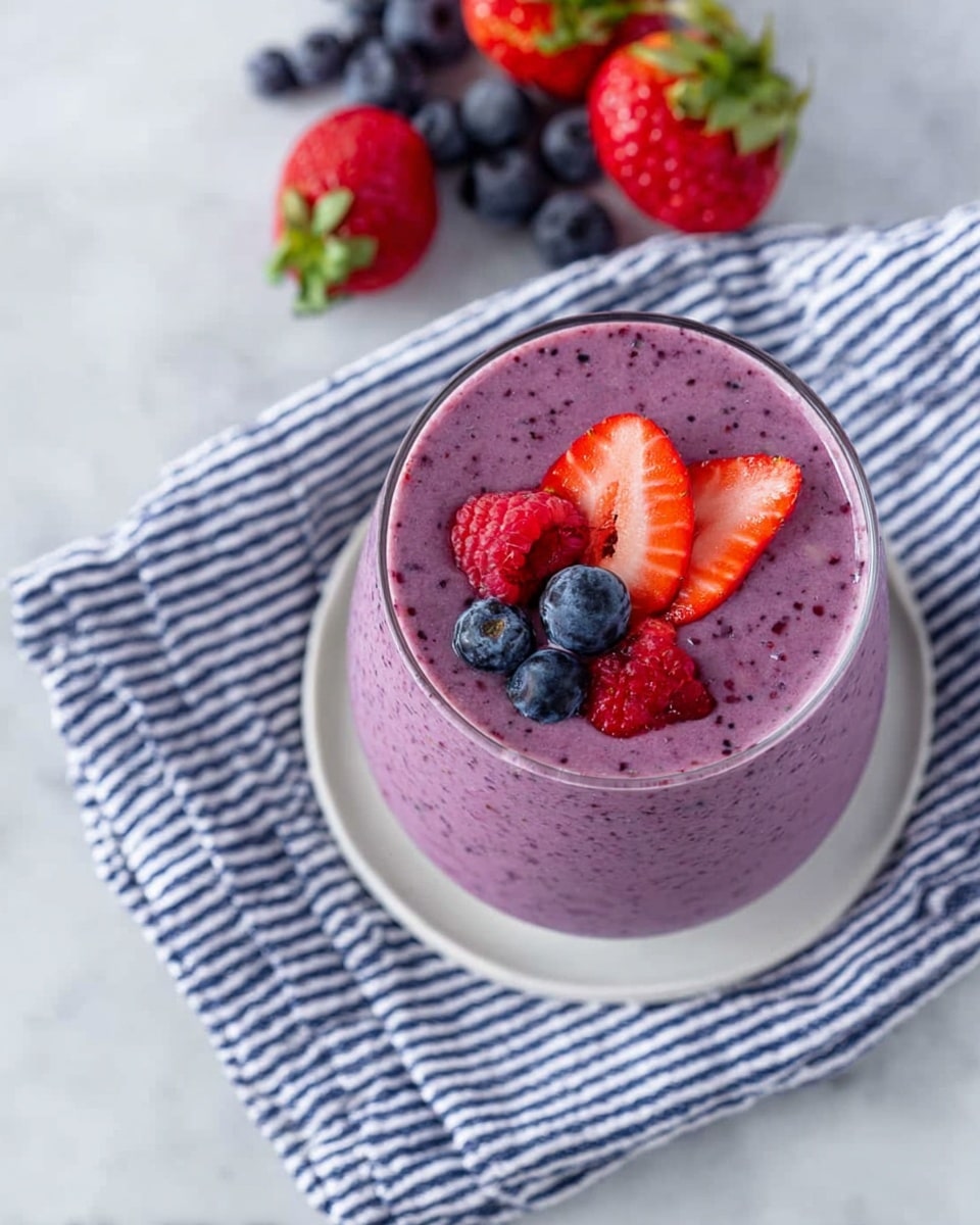 The image shows a glass filled with a thick purple smoothie topped with a small mix of fresh whole blueberries, raspberries, and sliced strawberries arranged near the center. The glass sits on a white plate with a blue and white striped cloth underneath, placed on a white marbled surface. In the background, there are whole strawberries and scattered blueberries giving a fresh and colorful look. The smoothie texture is smooth with small visible berry specks inside. photo taken with an iphone --ar 4:5 --v 7