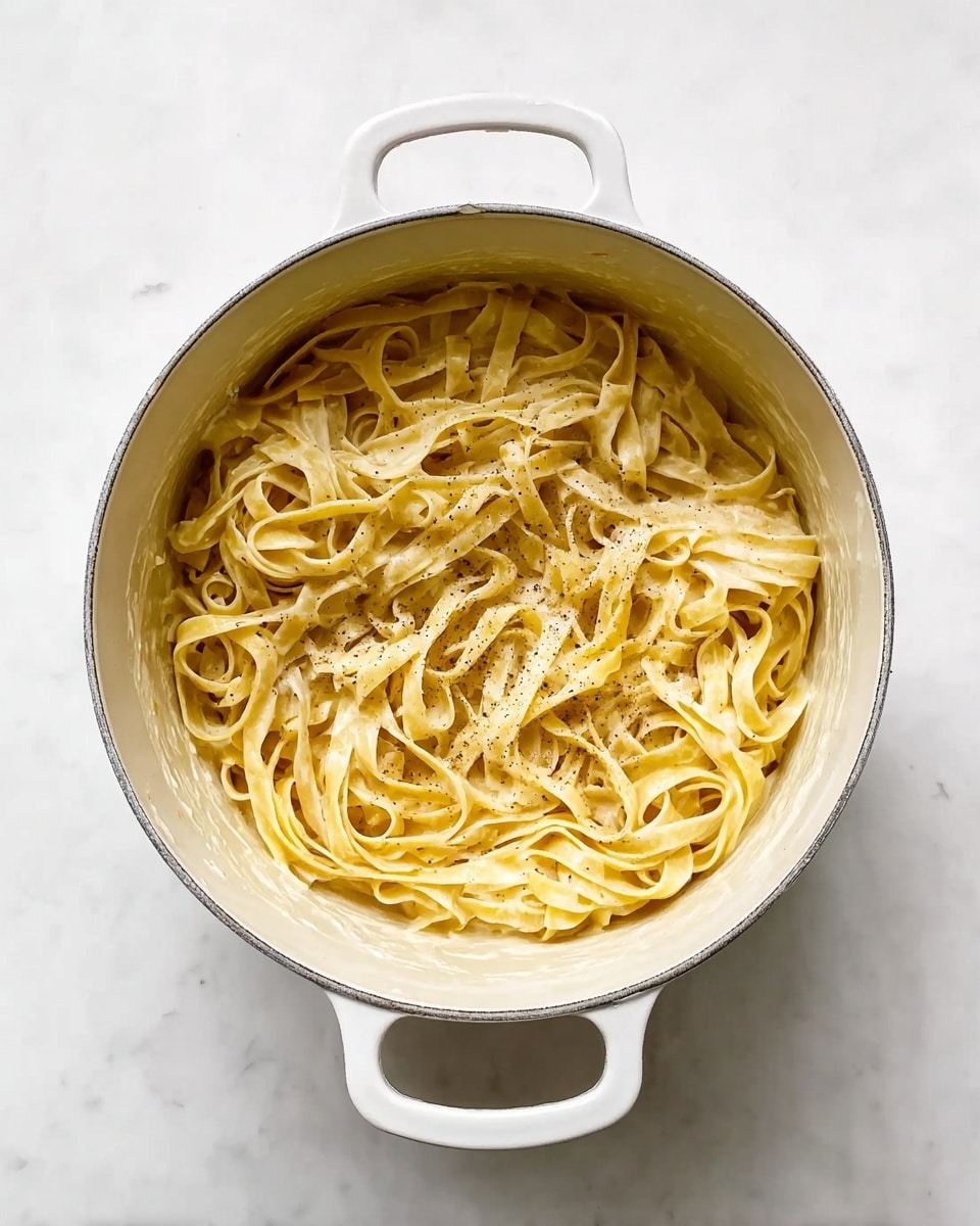 A white enamel pot filled with cooked fettuccine pasta, layered loosely and coated with a light cream-colored sauce with a few specks of black pepper sprinkled on top, all placed on a white marbled surface. Photo taken with an iphone --ar 4:5 --v 7