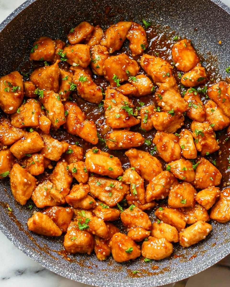 The image shows many small, golden-brown chicken pieces cooked in a textured gray non-stick pan, covered in a shiny brown sauce. The chicken pieces look tender and are sprinkled with small green herb leaves, adding a touch of color. The sauce is thick enough to coat the chicken evenly, giving a slightly sticky look. The pan is filled with these bite-sized chicken pieces, and the background is a white marbled surface. photo taken with an iphone --ar 4:5 --v 7