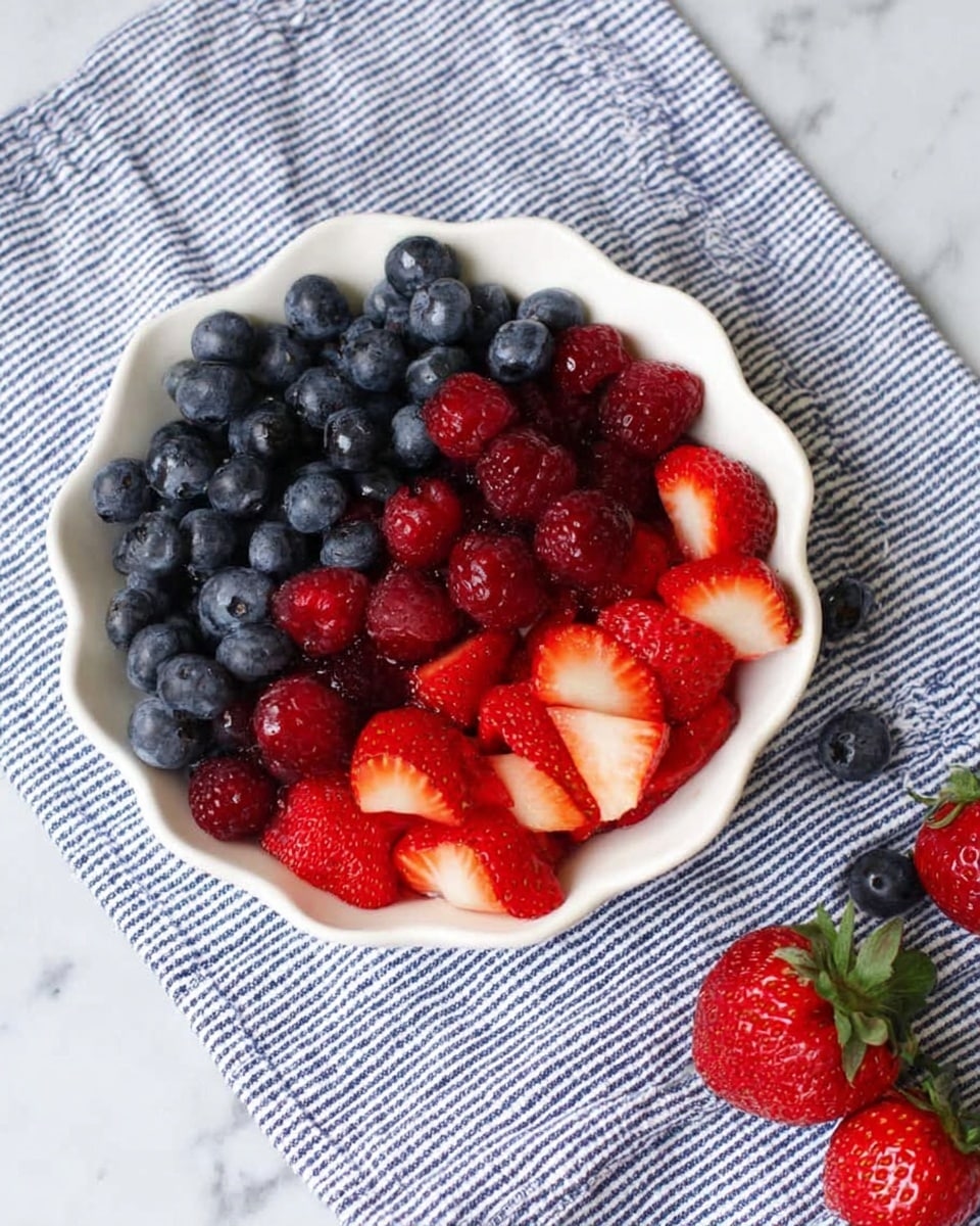 A white scalloped bowl filled with three layers of fresh berries: shiny, dark blue blueberries on top, bright red raspberries beneath them, and sliced strawberries with a red outside and white inside at the bottom. The bowl sits on a blue and white striped cloth on a white marbled surface, with a few whole strawberries and blueberries scattered beside the bowl. Photo taken with an iphone --ar 4:5 --v 7