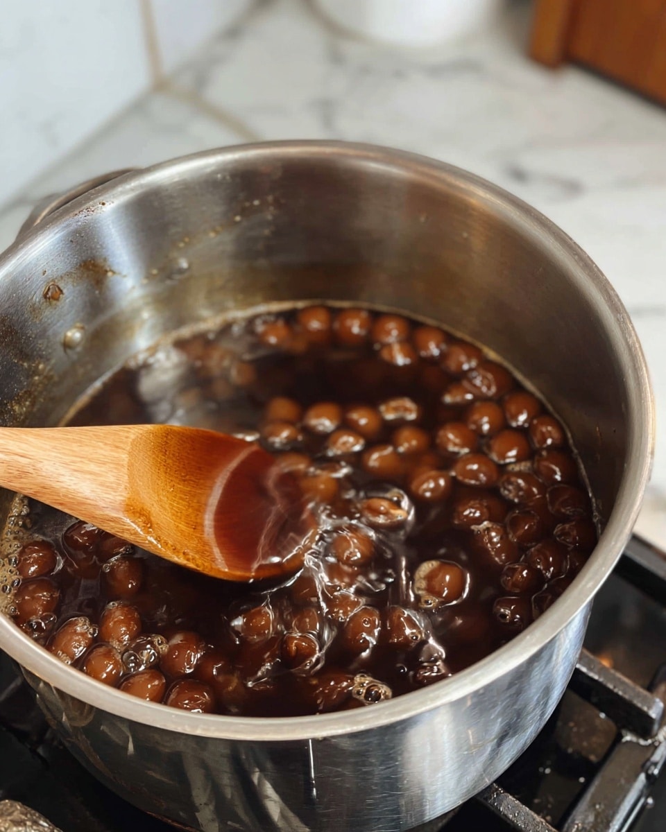 A close-up view of a stainless steel pot on a stove filled with small round brown pieces soaking in a thick dark brown liquid. A wooden spoon is stirring the mixture, showing the shiny and smooth texture of the liquid covering the pieces. The pot is set against a white marbled surface, and part of a white cabinet is visible in the background. photo taken with an iphone --ar 4:5 --v 7