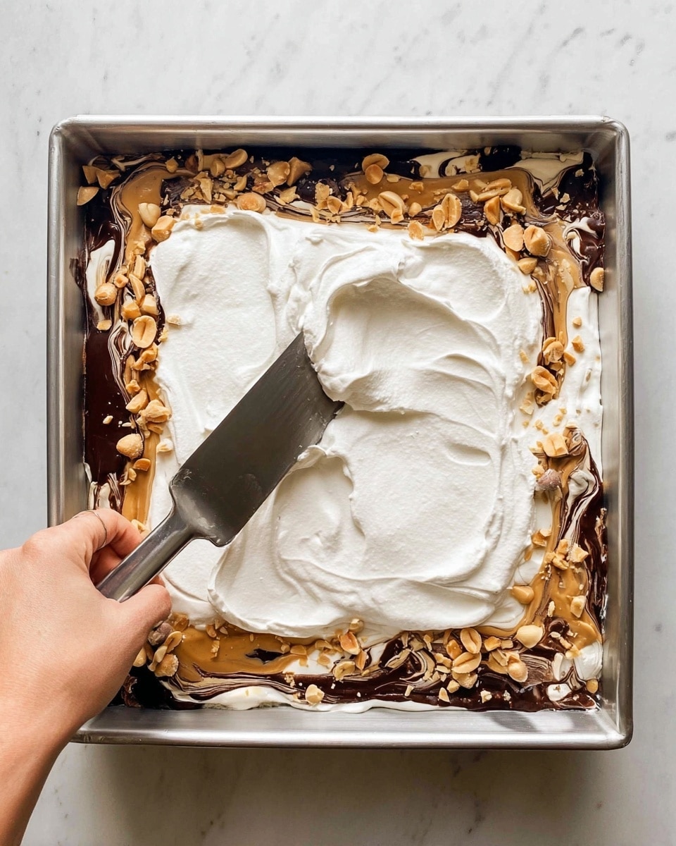 A square metal pan holds three visible layers of a dessert. The bottom layer is white and creamy, the middle layer has swirls of dark chocolate and light brown peanut butter mixed with scattered roasted peanuts, and the top layer is a thick, smooth spread of white whipped cream being spread evenly with a metal spatula held by a woman's hand. The pan rests on a white marbled surface. photo taken with an iphone --ar 4:5 --v 7
