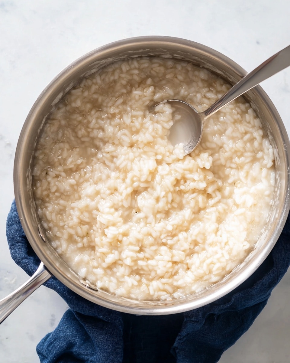 A shiny steel pot filled with creamy, light beige risotto that has a soft, thick texture and plump rice grains visible throughout. The risotto is partially stirred, showing a small curved space where a silver spoon with a smooth handle is placed inside. The pot sits on a dark blue cloth, all set against a white marbled surface. photo taken with an iphone --ar 4:5 --v 7