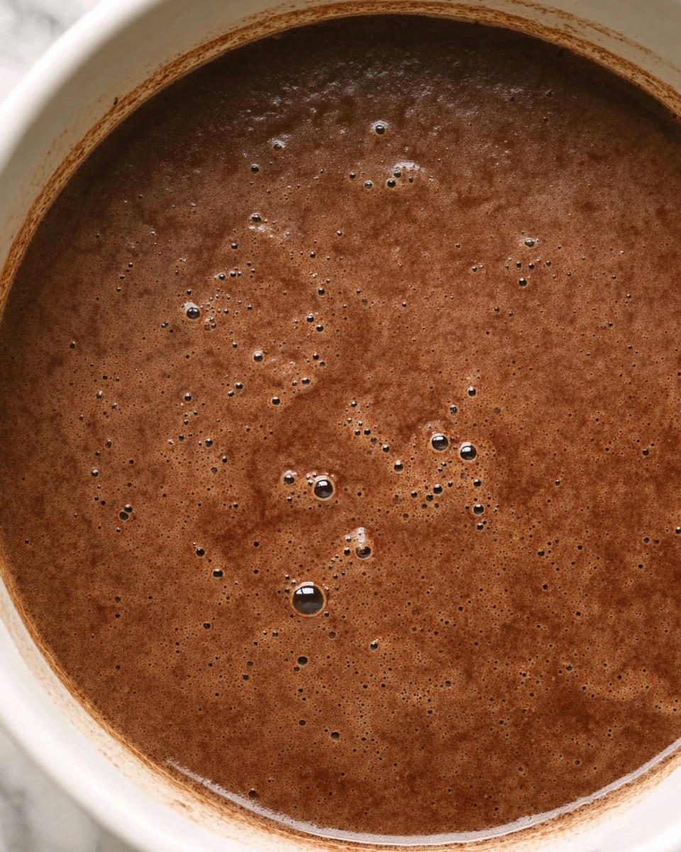 The image shows a close-up of a single layer of thick, dark brown liquid with small bubbles on the surface, cooking inside a white pot. The liquid has a slightly grainy texture with varying shades of brown and darker specks scattered throughout. The edges of the pot are visible, showing the contrast between the smooth white interior and the rich chocolatey liquid inside. The surface beneath the pot appears as a white marbled texture. photo taken with an iphone --ar 4:5 --v 7