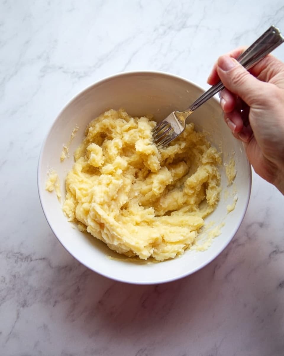 A white bowl holds a soft, pale yellow mashed mixture with a slightly lumpy texture. A woman’s hand is using a metal fork to mash the contents inside the bowl, which rests on a white marbled surface. The lighting is natural and bright, showing the smooth yet slightly chunky consistency of the food inside the bowl. photo taken with an iphone --ar 4:5 --v 7