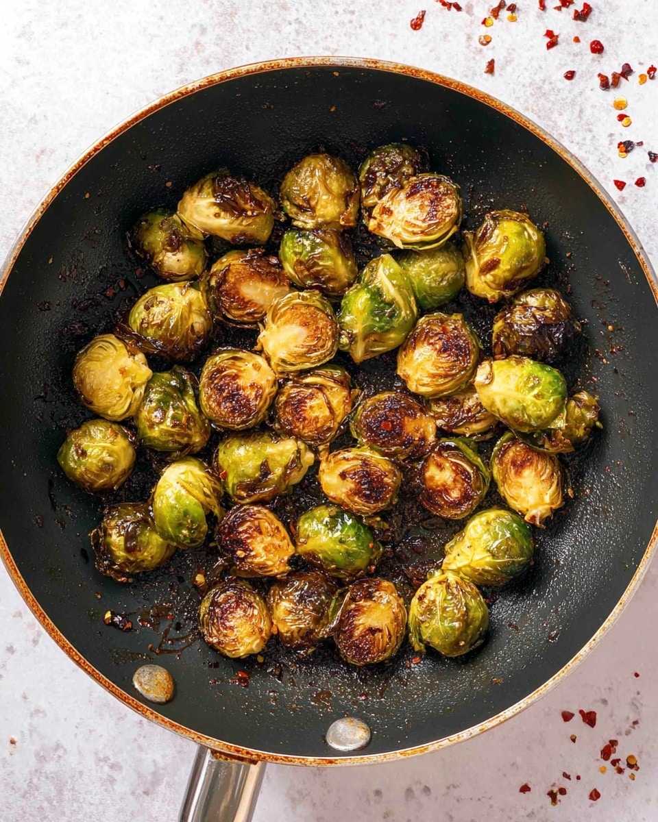 The image shows a black frying pan filled with roasted Brussels sprouts. The Brussels sprouts are golden brown with some darker charred spots, giving a crispy texture. They are small to medium-sized, some whole and some opened slightly, with uneven roasting marks. The pan has a shiny, oily surface with bits of cooked residue and the handle is visible at the bottom right. The background is a white marbled texture scattered lightly with red chili flakes. photo taken with an iphone --ar 4:5 --v 7