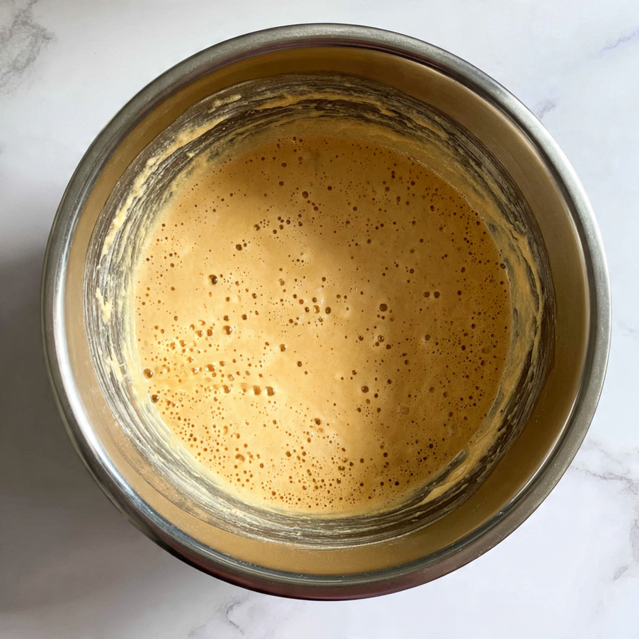 The image shows a top view of a metallic mixing bowl with a layer of bubbly, light brown batter inside. The batter looks smooth and slightly thick with some small air bubbles on the surface, giving it a frothy texture. The inner walls of the bowl have some batter residue clinging to them. The bowl sits on a white marbled surface. photo taken with an iphone --ar 4:5 --v 7