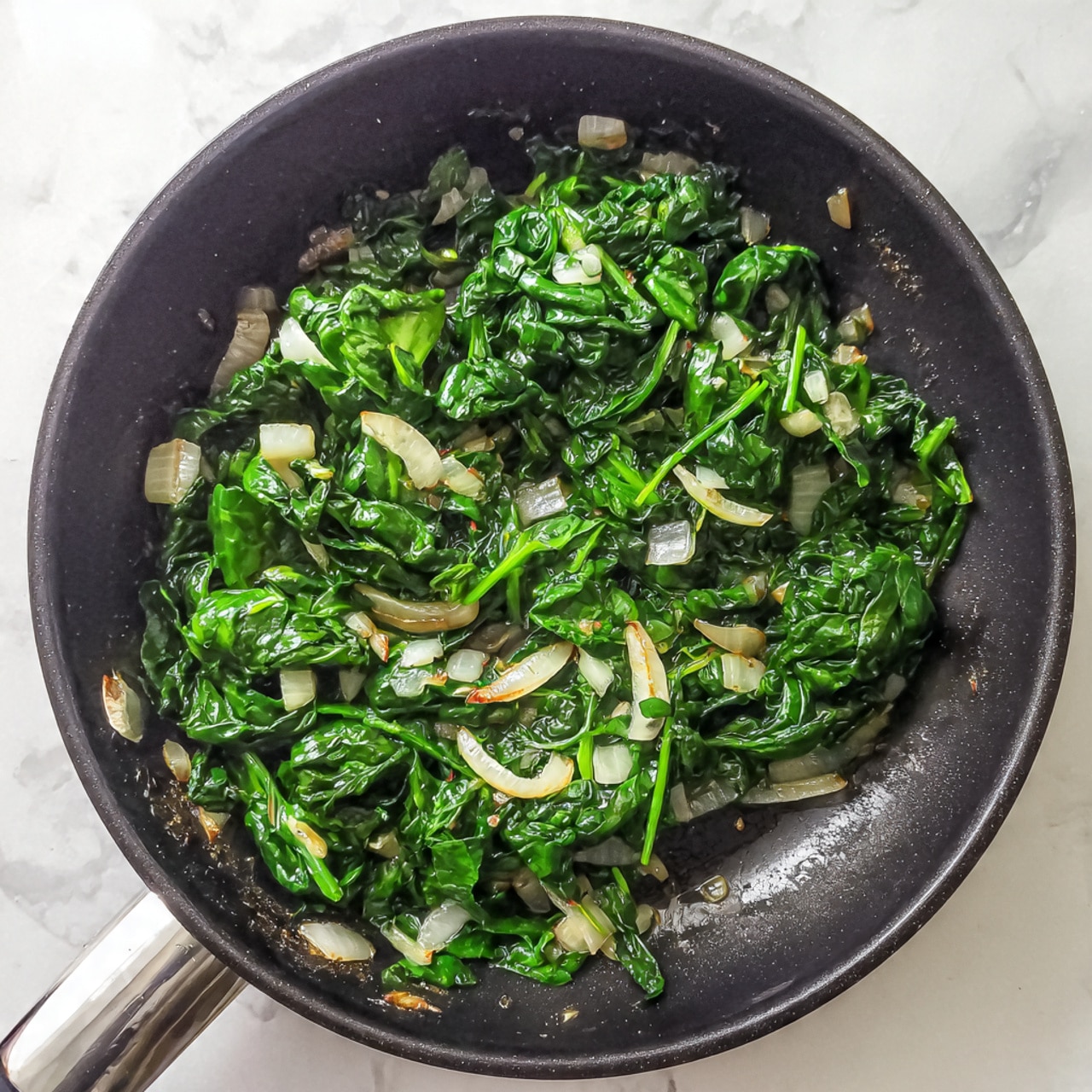 The image shows a close-up of cooked spinach and chopped white onions in a black frying pan. The spinach is bright green with a slightly wilted texture, and the onions are soft and translucent with a light cream color. The pan has some shiny spots where the cooking oil reflects light. The pan rests on a white marbled surface. photo taken with an iphone --ar 4:5 --v 7