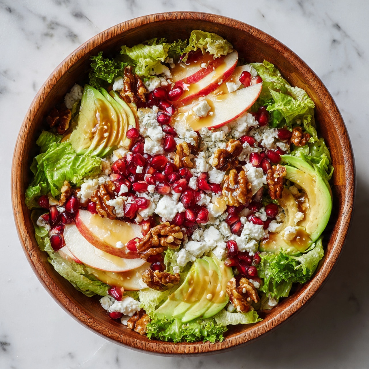 This dish is a fresh salad served in a wooden bowl, filled with many layers. The bottom layer is bright green curly lettuce leaves spread all over. Scattered on top are dark red pomegranate seeds, creamy white and blue chunks of cheese, light green avocado slices, and small brown walnut halves. There are also thin slices of pale yellow apple peeking through the greens. Everything is drizzled with a shiny golden dressing that adds a wet look. The bowl and food are photographed on a white marbled surface. photo taken with an iphone --ar 4:5 --v 7