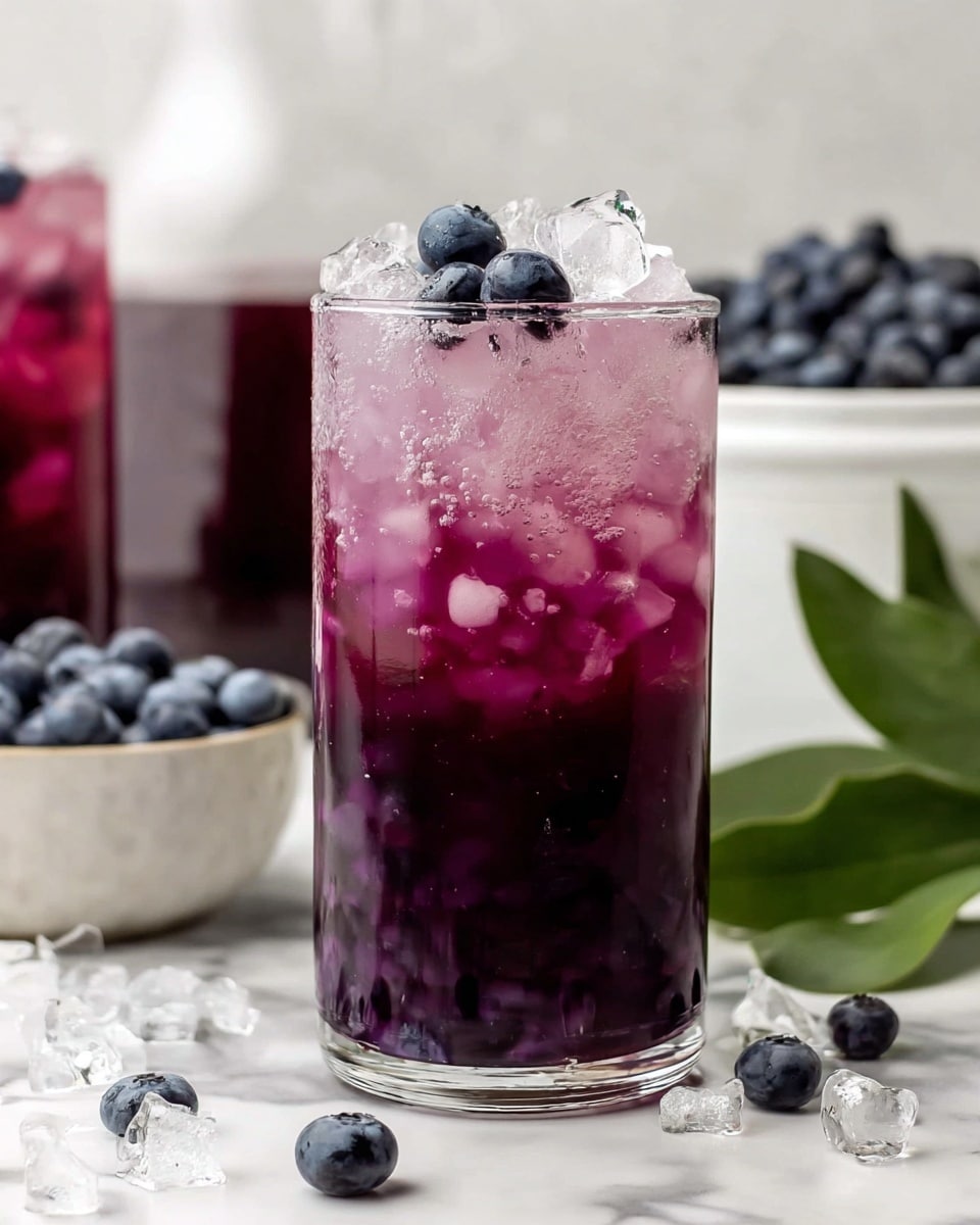 A tall clear glass filled with three layers: the bottom layer is a dark purple liquid, the middle layer is crushed ice mixed with a lighter purple liquid, and the top layer has a few whole blueberries resting on the ice. The glass is placed on a white marbled surface scattered with loose blueberries and ice pieces. In the background, there is a white bowl filled with blueberries and another taller glass with a similar purple drink. A green leafy sprig is to the right side, near the glass. Photo taken with an iphone --ar 4:5 --v 7