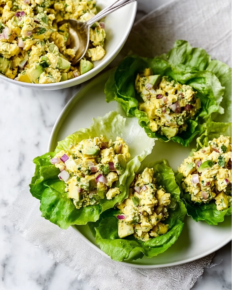 The image shows a white plate on a white marbled surface holding bright green leafy lettuce cups filled with a chunky mixture. The mixture inside the lettuce cups is made of pale yellow chopped eggs mixed with small pieces of green avocado and diced red onions, adding splashes of purple. The lettuce leaves have a crisp, curly texture and form small bowls for the filling, which looks creamy yet chunky. Behind the lettuce cups is a round white bowl filled with the same egg and avocado mixture, with a spoon resting inside. The whole scene has natural light, highlighting the fresh colors and textures. Photo taken with an iphone --ar 4:5 --v 7