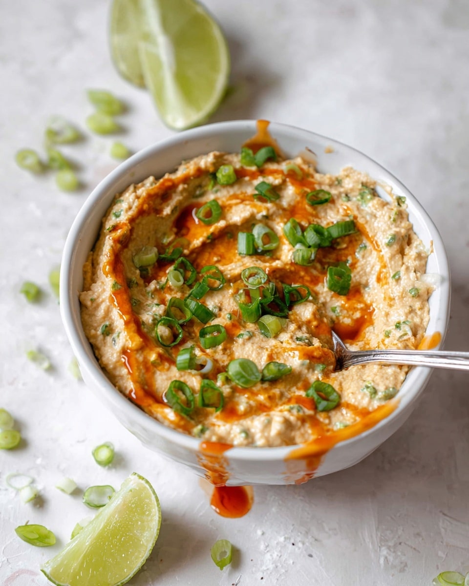A white bowl filled with a creamy textured dip that has a light beige color mixed with some specks of green herbs, swirled on top with a drizzle of reddish-orange sauce that slightly drips down the side of the bowl. On top of the dip, there are bright green chopped scallions scattered evenly, while a silver spoon rests inside the bowl on the right side. Around the bowl, there are a few slices of scallion and two lime wedges in the background, all placed on a white marbled surface. photo taken with an iphone --ar 4:5 --v 7