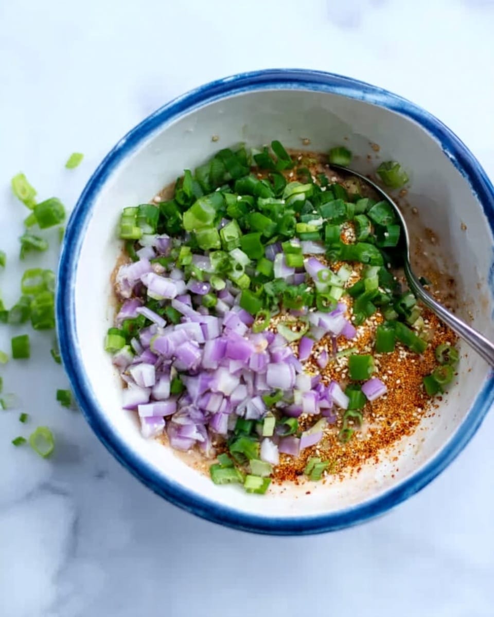 The image shows a white bowl with a blue inside, sitting on a white marbled surface. Inside the bowl, the base layer is light brown textured with tiny red flecks, likely a seasoning or sauce. On top of this base are chopped green onions and thin slices of light purple shallots, creating a mix of green and purple colors concentrated in the center. A small part of a metal spoon is resting inside the bowl on the left side. Scattered chopped green onions are visible on the surface near the bowl. Photo taken with an iphone --ar 4:5 --v 7