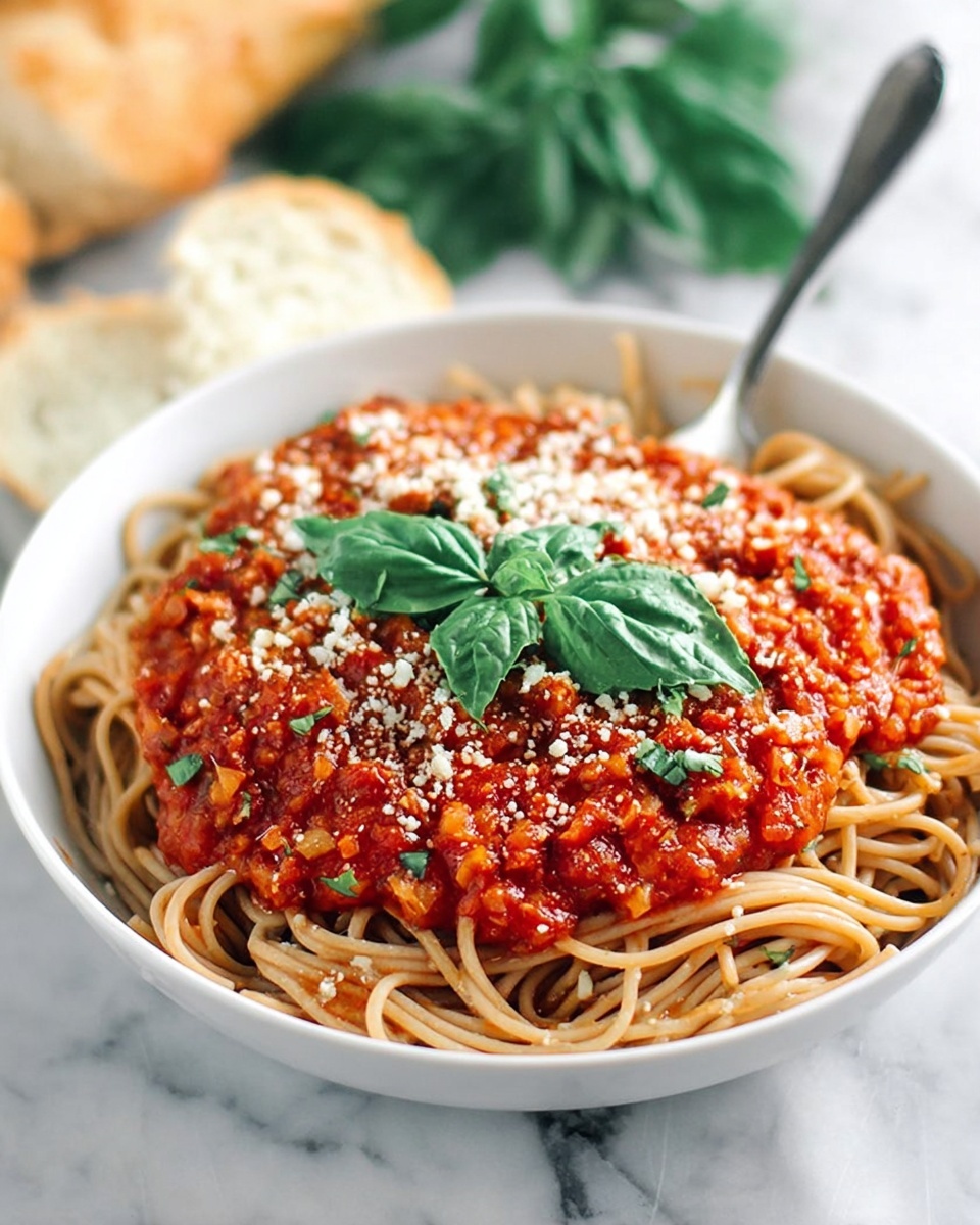 A white bowl filled with a bed of light brown spaghetti noodles at the bottom layer, topped with a thick, chunky red tomato sauce mixed with small pieces of vegetables and herbs. The sauce has a rich texture and a sprinkle of grated white cheese on top. Fresh green basil leaves are placed in the center of the sauce. A silver fork is stuck into the noodles at the back left side of the bowl. The background is a white marbled surface with some fresh basil leaves and a piece of bread blurred in the distance. Photo taken with an iphone --ar 4:5 --v 7