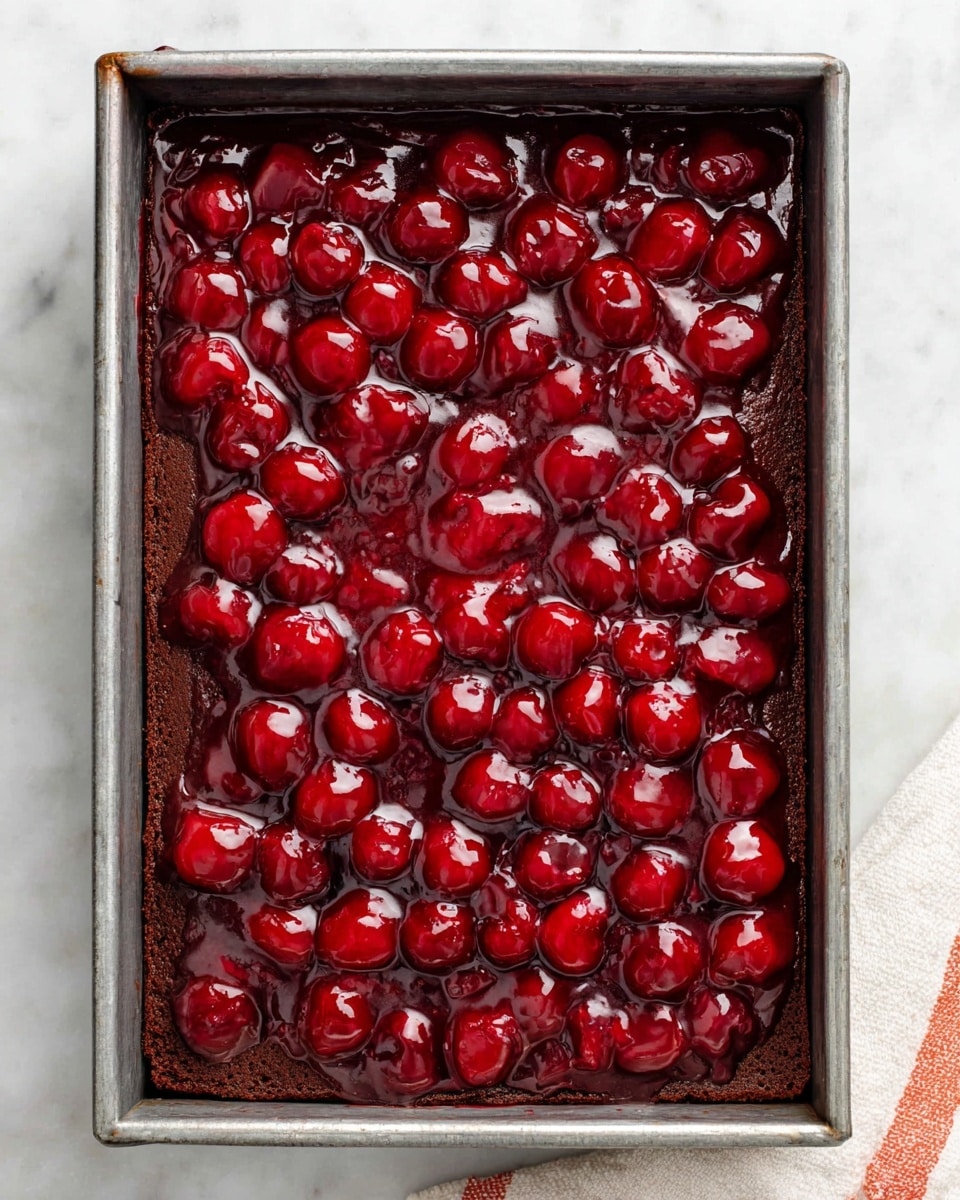A rectangular metal baking pan filled with a dark brown cake layer at the bottom, covered by a thick, shiny, bright red cherry topping with whole cherries spread evenly across the surface. The cherries look plump and glossy, with the red topping slightly uneven in texture and reflecting light. The pan rests on a white marbled surface, with a small corner of a white cloth with orange stripes visible at the bottom right. photo taken with an iphone --ar 4:5 --v 7