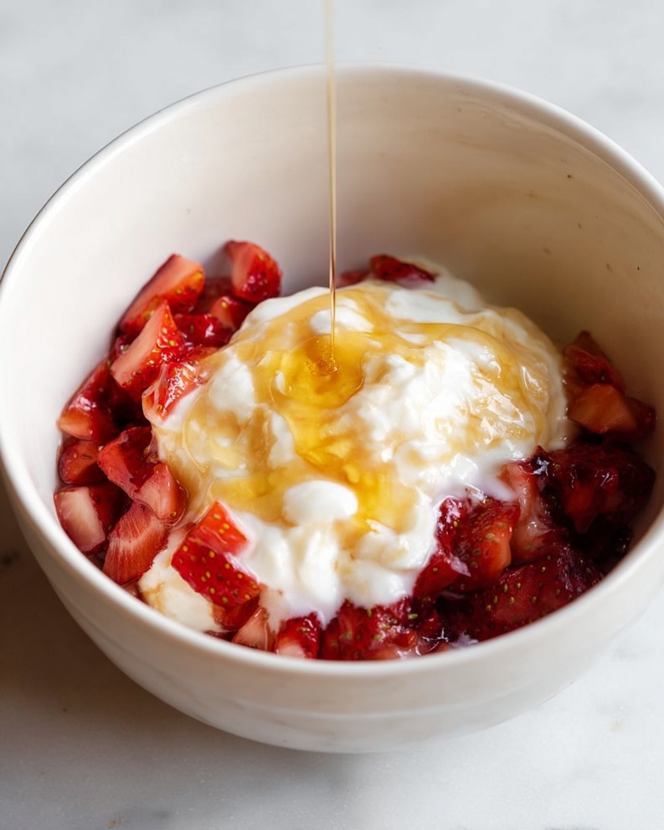 The image shows a white bowl placed on a white marbled surface, filled with three main layers. The bottom layer is made of small, finely chopped pieces of red strawberries, giving a fresh and juicy look. The middle layer is a beige creamy mixture that looks smooth and thick. On top, there is a dollop of white whipped cream, which looks light and fluffy, being covered by a glossy, golden drizzle of honey slowly dripping from above into the bowl. photo taken with an iphone --ar 4:5 --v 7