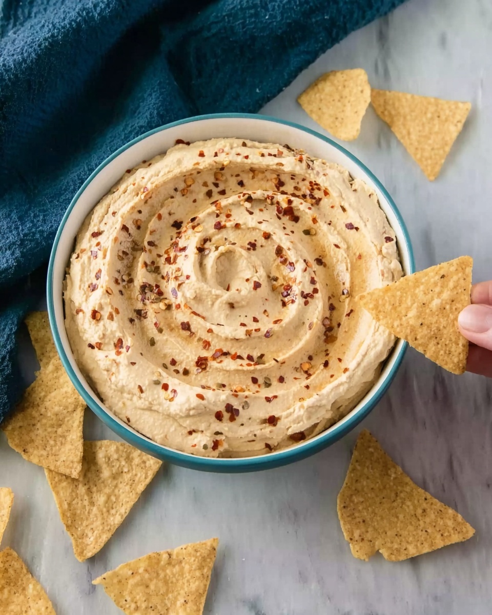 A white bowl filled with creamy hummus, spread with light swirls and sprinkled with red chili flakes on top. The hummus has a smooth texture with tiny visible bits. Around the bowl, there are several light beige triangular tortilla chips scattered on a white marbled surface. A woman's hand is holding one tortilla chip, dipping it into the hummus near the edge of the bowl. A dark blue cloth is partially visible in the upper left corner. Photo taken with an iphone --ar 4:5 --v 7