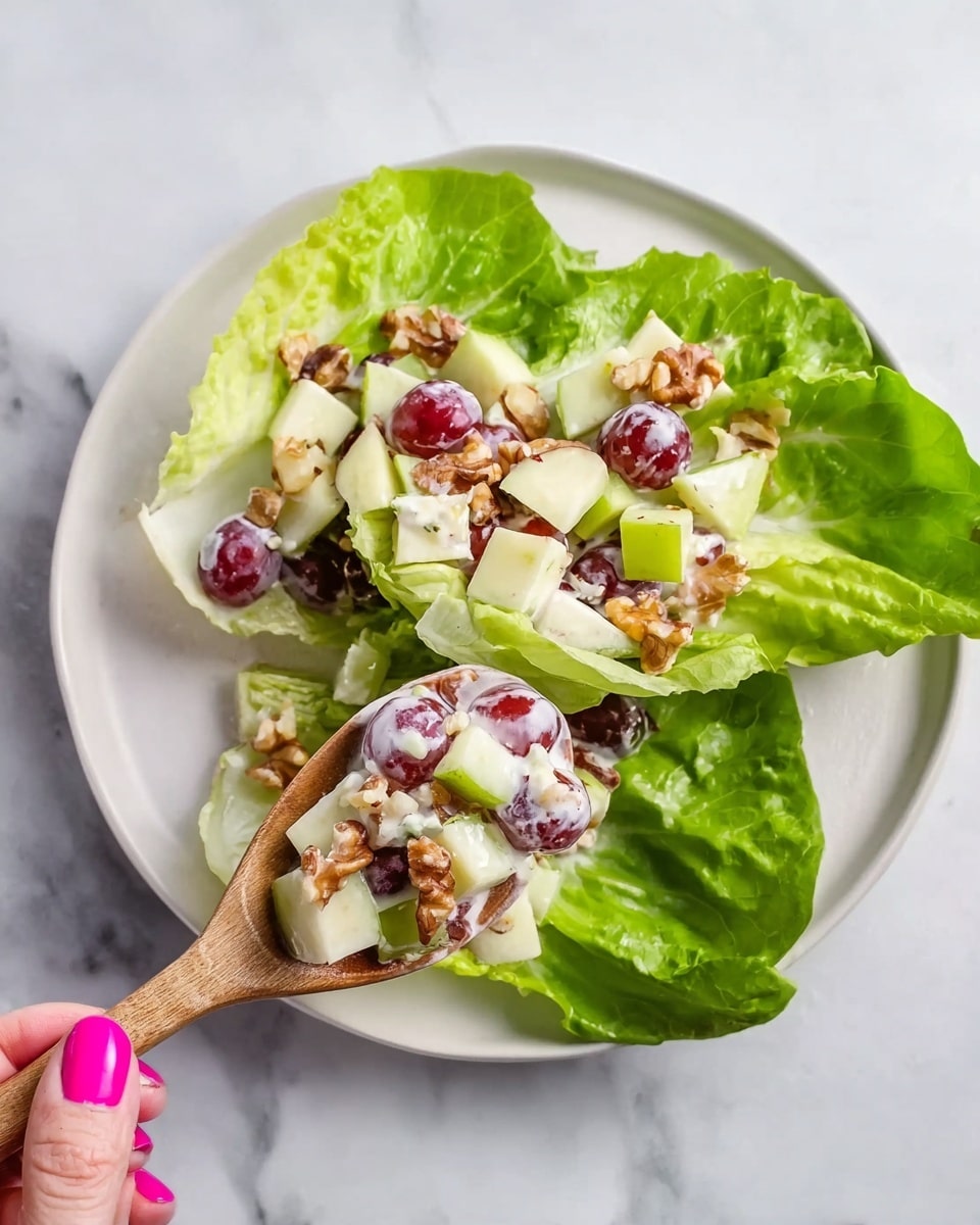 A white plate sits on a white marbled surface with fresh green butter lettuce leaves as the base layer, showing a glossy and smooth texture. On top, there is a mix of chopped light yellow apples, red grapes, and beige walnut pieces, all coated with a creamy white dressing. A wooden spoon with a bright pink nail on a woman's hand holds a serving of this fruity salad over the leaves. The colors are fresh and vibrant with contrasting greens, reds, and creamy whites, making the dish look fresh and healthy photo taken with an iphone --ar 4:5 --v 7