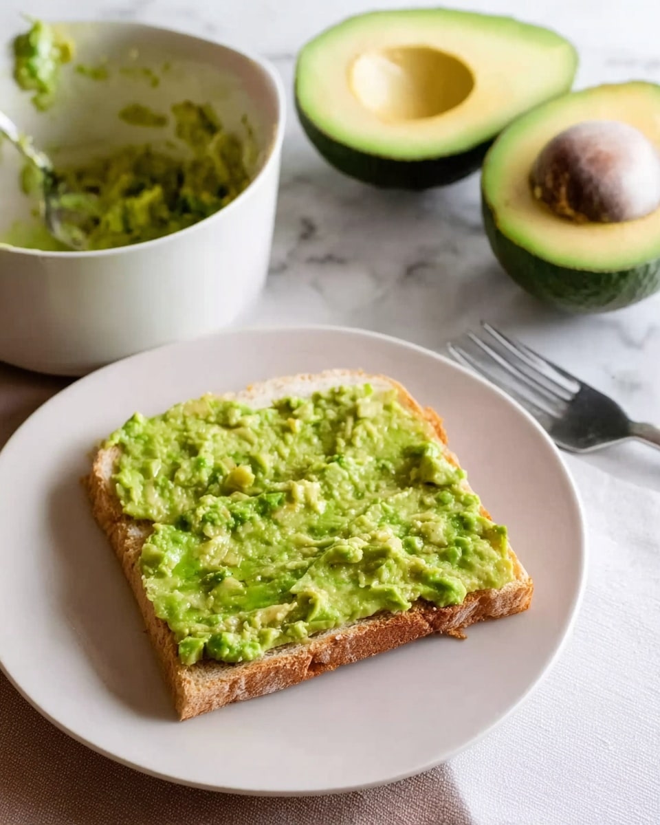 A close-up view shows a slice of square bread on a white plate, topped with a thick, uneven layer of mashed green avocado, with some small chunks visible. Next to the plate is a white bowl containing leftover mashed avocado and a fork resting inside it. Behind the bowl, there is a halved avocado with a large brown seed in the center on a white marbled surface. photo taken with an iphone --ar 4:5 --v 7