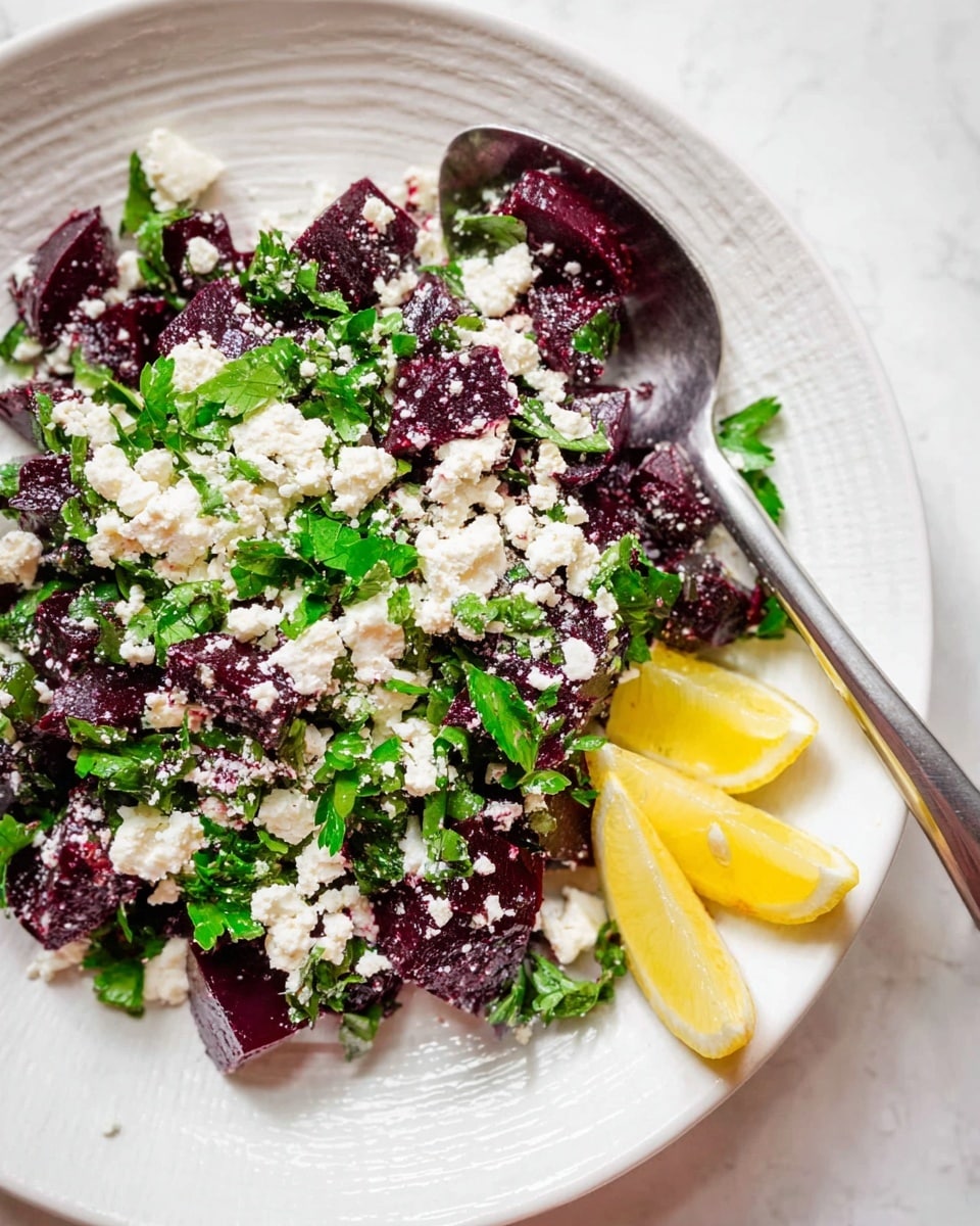 A white plate with a salad made of deep purple beet pieces scattered throughout, topped with white crumbled cheese that covers the beets and some of the plate, mixed with chopped bright green leafy herbs spread evenly on top and around. Two yellow lemon wedges rest on the edge of the plate. A large silver spoon sits on the right side of the plate, partially under the salad. The plate is on a white marbled surface with subtle grooves radiating from the center photo taken with an iphone --ar 4:5 --v 7