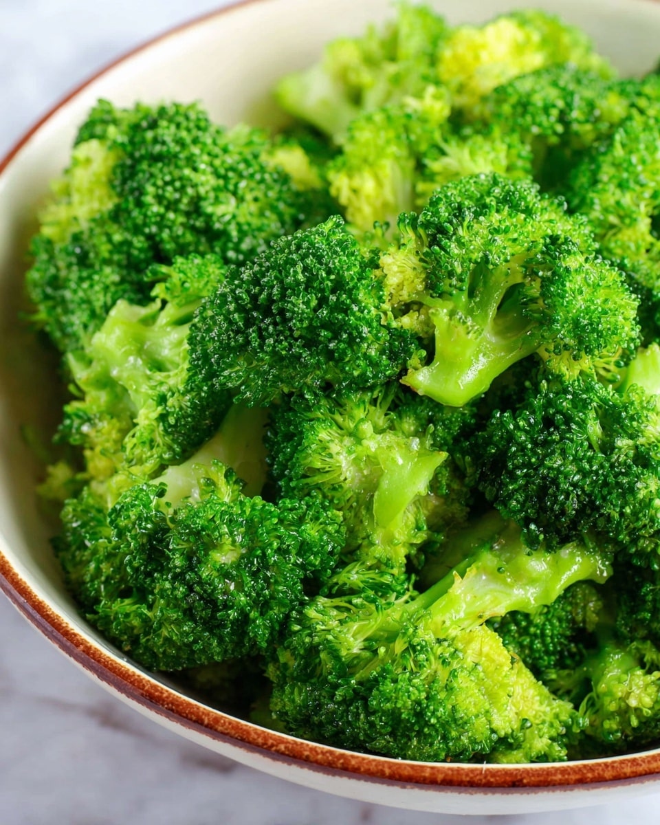 A close-up of a bowl filled with bright green broccoli florets, showing the detailed texture of the small buds and stems. The broccoli is fresh with a slight shine, piled up inside a white bowl with a brown rim. The scene is set on a white marbled surface, highlighting the vibrant green color and the moist texture of the broccoli. photo taken with an iphone --ar 4:5 --v 7