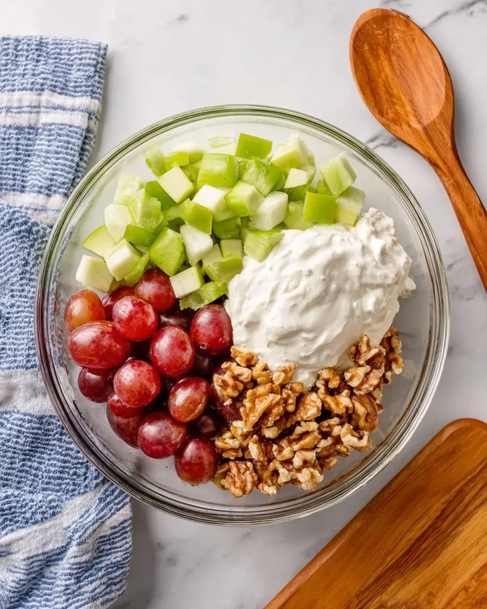 The image shows a clear glass bowl on a white marbled surface filled with five distinct layers: chopped green celery on the top left, chopped light green apple on the top right, a dollop of thick white cream or yogurt in the center, halves of red grapes on the bottom left, and chopped brown walnuts on the bottom right. To the top right of the bowl, there is a wooden spoon, and on the top left, a blue and white striped cloth. At the bottom left, a wooden cutting board is partially visible. photo taken with an iphone --ar 4:5 --v 7