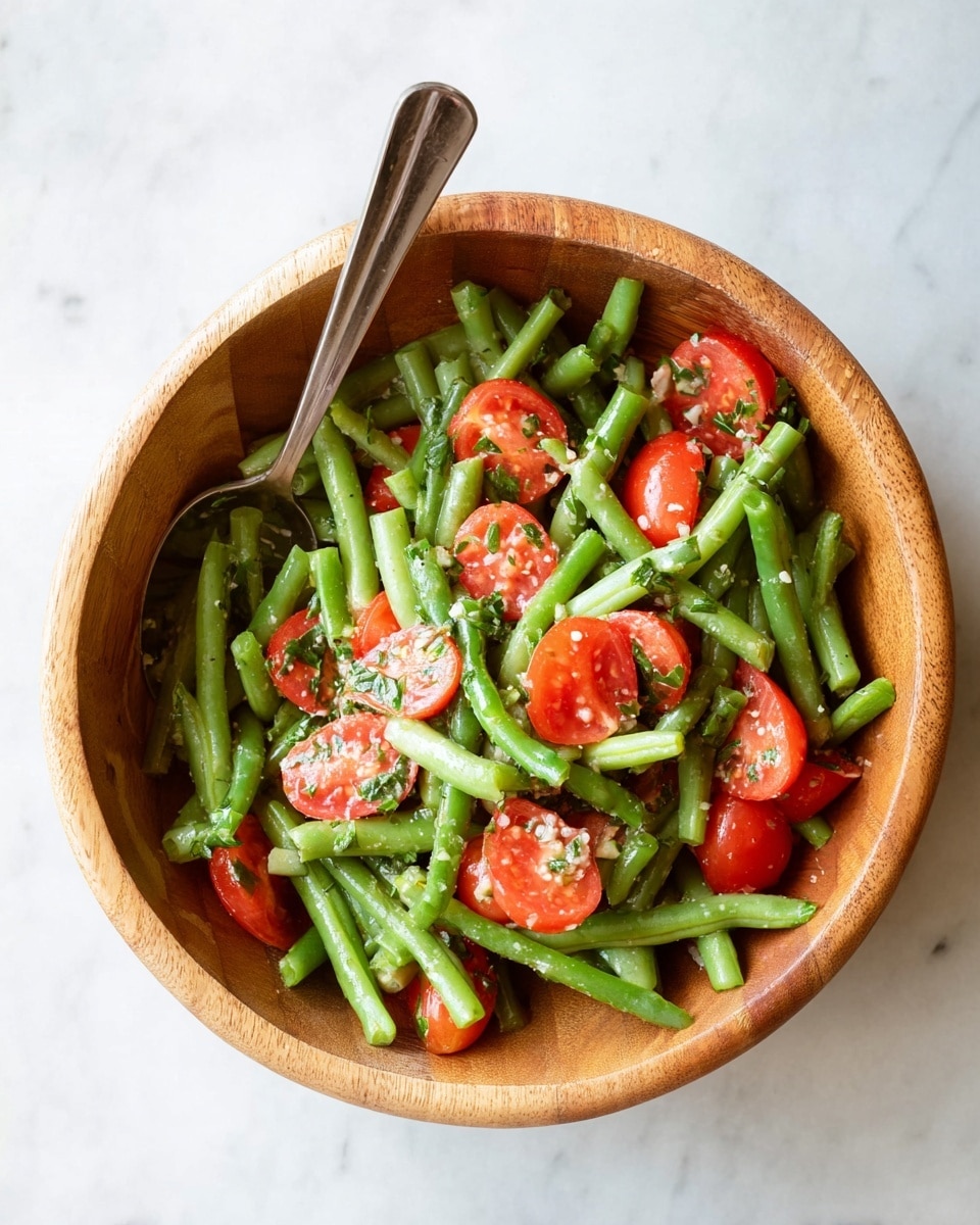 A wooden bowl holds a fresh salad made of bright green pieces of green beans layered with sliced red tomatoes scattered evenly throughout. The salad is topped with small bits of chopped green herbs and a light sprinkle of white seasoning. A shiny metal serving spoon rests inside the bowl on the left side, with its spoon part touching some tomatoes and beans. The bowl sits on a white marbled surface. photo taken with an iphone --ar 4:5 --v 7