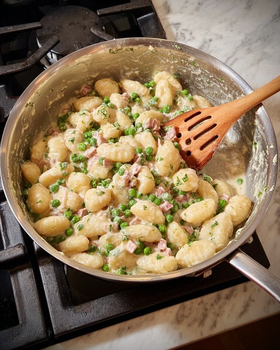 This image shows a stainless steel pan filled with a creamy gnocchi dish on a stove. The dish has three main visible layers: soft, plump, light beige gnocchi pieces forming the base; small, vibrant green peas scattered evenly throughout; and small chunks of pale pink meat mixed in sparsely. The creamy sauce coats the gnocchi and peas, giving a smooth, slightly shiny texture. A wooden spoon with holes is resting in the pan, mixing the ingredients. The pan is placed on a gas stove with black grates, and the counter around is a white marbled texture. photo taken with an iphone --ar 4:5 --v 7
