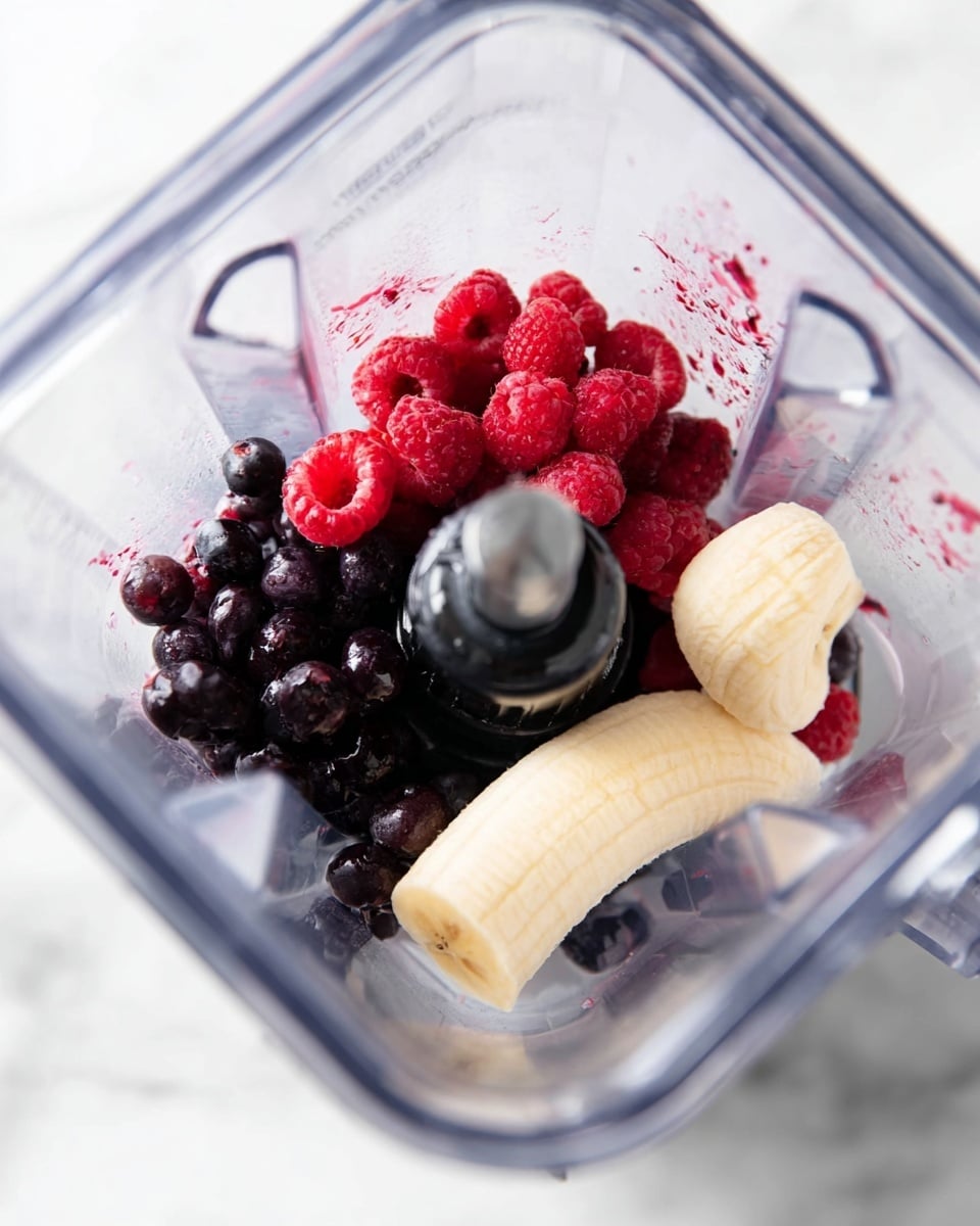 Inside a clear blender container, there is a layer of dark purple blueberries at the bottom left, mixed with bright red raspberries stacked slightly above and to the right. On the right side, a peeled section of banana with a soft yellow color rests next to the berries. The blender blade and center rod are visible in the middle, surrounded by the fruit, all set on a white marbled surface. Photo taken with an iphone --ar 4:5 --v 7