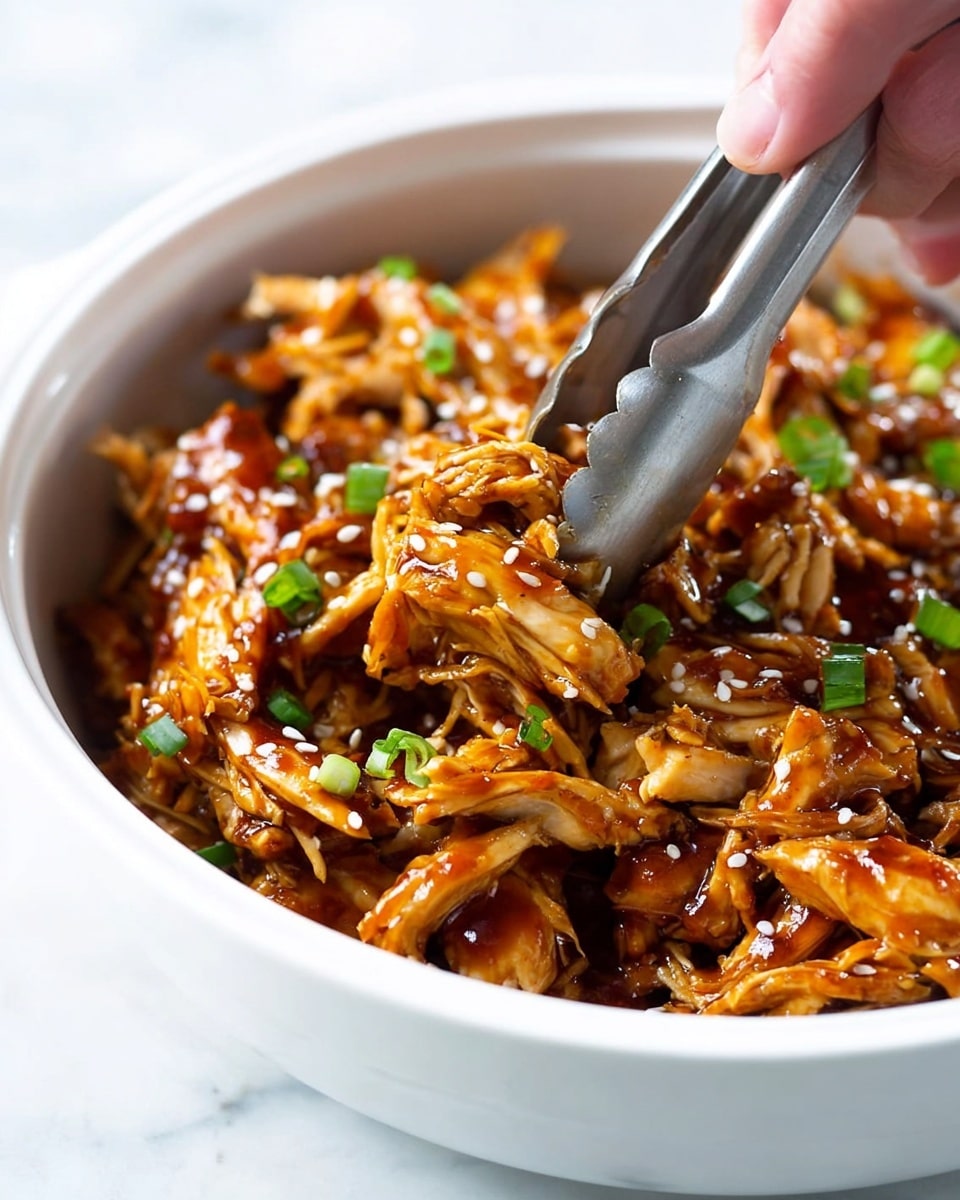 A close-up view of a white bowl filled with shredded cooked chicken pieces covered in a shiny, dark orange-brown sauce. The chicken is sprinkled with small white sesame seeds and some chopped green onions. A pair of metal tongs held by a woman's hand is picking up some of the chicken from the bowl. The bowl is placed on a white marbled surface. photo taken with an iphone --ar 4:5 --v 7