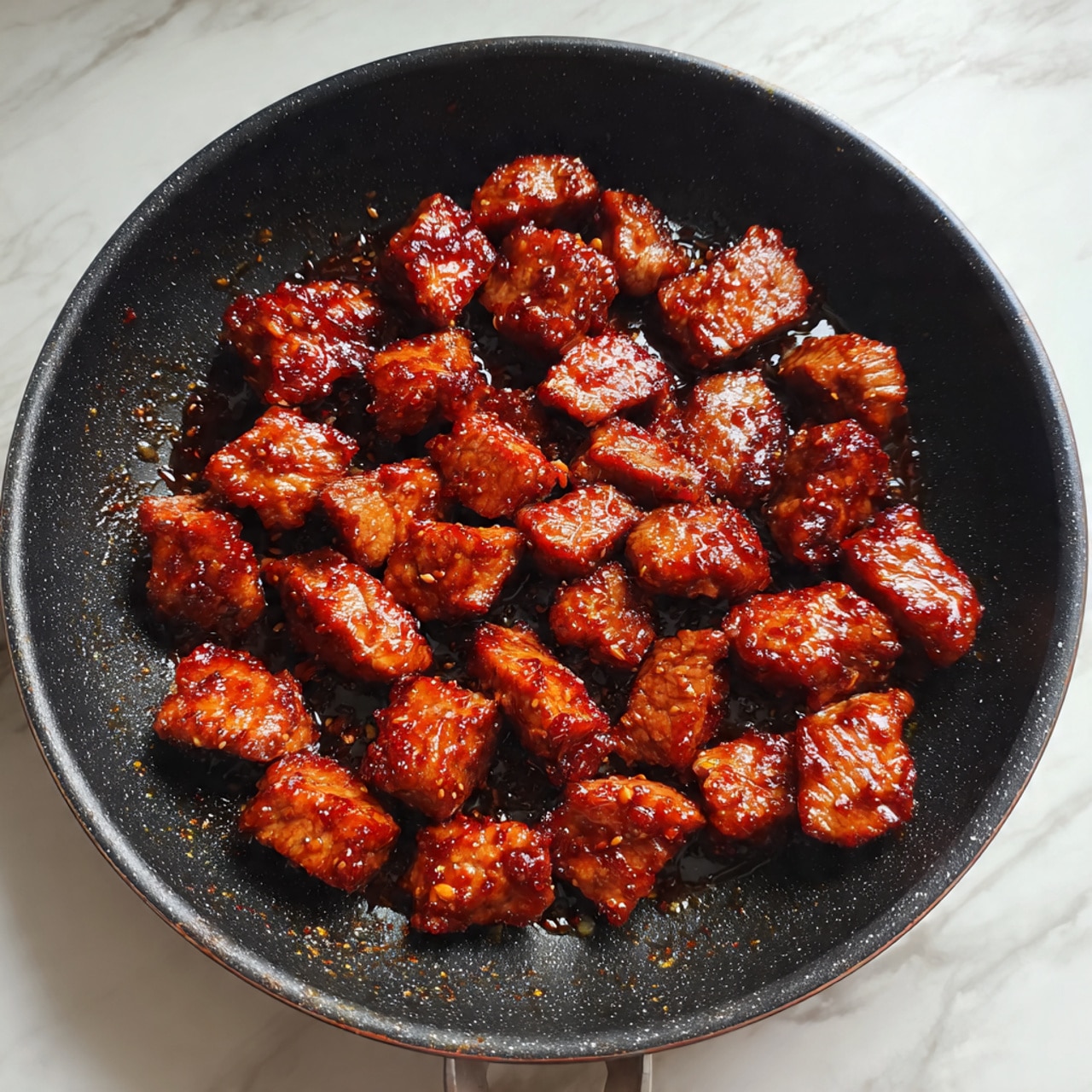 A close-up view of a frying pan filled with multiple pieces of cooked meat, rich brown in color with a slightly shiny, textured surface indicating a savory sauce or marinade. The meat pieces vary in size and shape, scattered evenly across the dark pan's circular base, sitting on a white marbled surface. The texture of the meat looks tender and juicy with visible slight caramelization on some edges. Photo taken with an iphone --ar 4:5 --v 7