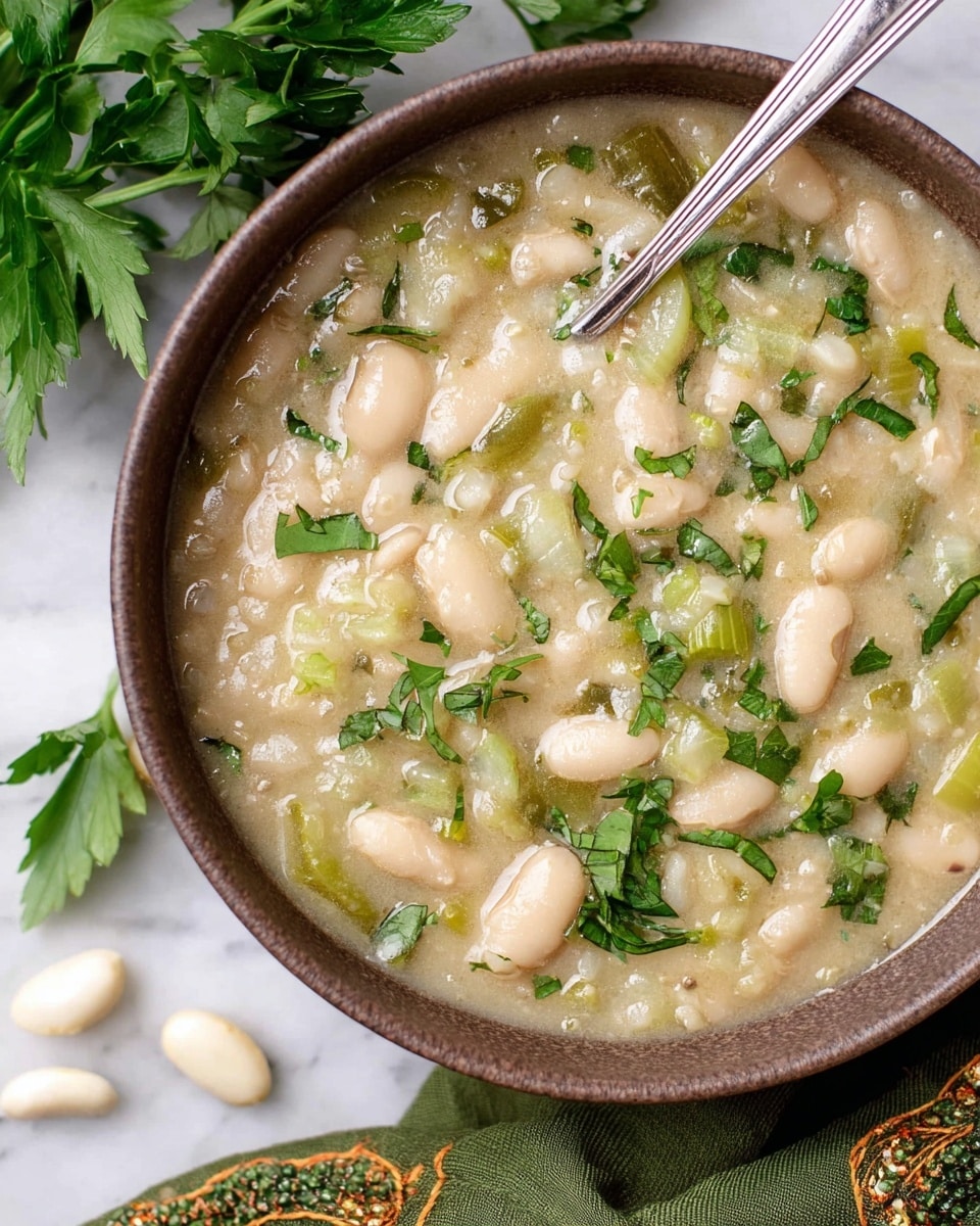 A close-up view of a brown bowl filled with thick white bean soup that has a creamy texture. The soup shows many white beans and small pieces of green celery mixed in a pale creamy broth. Fresh green herbs, roughly chopped, are sprinkled on top, adding a pop of color across the surface. A silver spoon rests inside the bowl, with its handle angled to the right. The bowl sits on a white marbled surface, with some fresh green parsley leaves and a few loose white beans scattered around. A green cloth with a decorative border is draped at the edge, partly under the bowl. Photo taken with an iphone --ar 4:5 --v 7