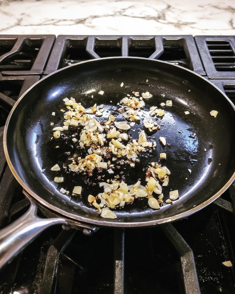 A black frying pan with a shiny surface sits on a gas stove with black grates. Inside the pan, small pieces of light yellow and white chopped garlic are scattered and lightly cooked, showing some browning spots. The pan handle extends to the left and the whole scene is on a white marbled surface. Photo taken with an iphone --ar 4:5 --v 7