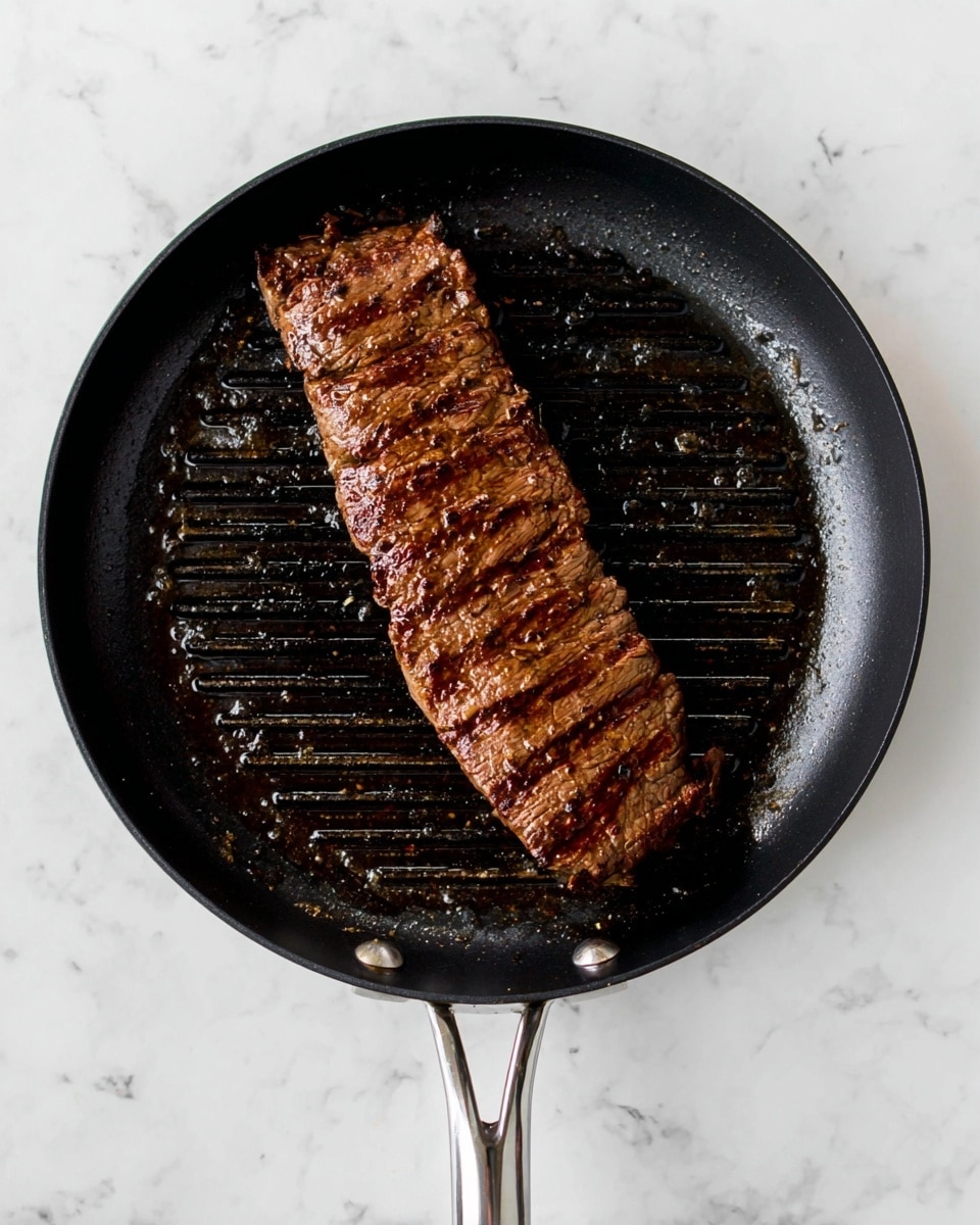 A single long piece of cooked meat with dark brown grill marks lies in the center of a black grill pan with a metal handle, resting on a white marbled surface. The meat shows a textured surface with seasoning specks and a slightly glossy finish from the cooking juices, and the pan has blackened oil spots and grill ridges clearly visible beneath the meat. Photo taken with an iphone --ar 4:5 --v 7
